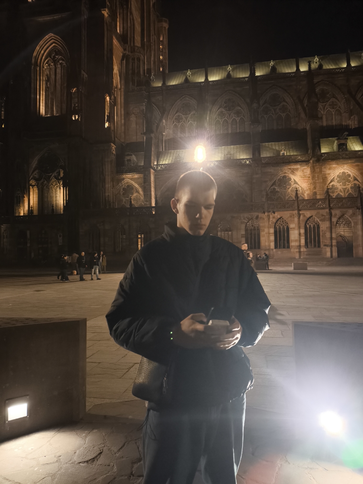 A low-angle shot at night features a male figure standing in front of Strasbourg Cathedral in Strasbourg, France. The cathedral's detailed Gothic facade forms the backdrop, illuminated with warm light. The male subject is looking down at a cell phone in his hands, wearing a dark jacket and dark pants. There is a black shoulder bag behind him. Light sources are visible in the foreground and above the man's head, causing glare. Several people are blurred in the background. The ground is stone-paved and unevenly lit.