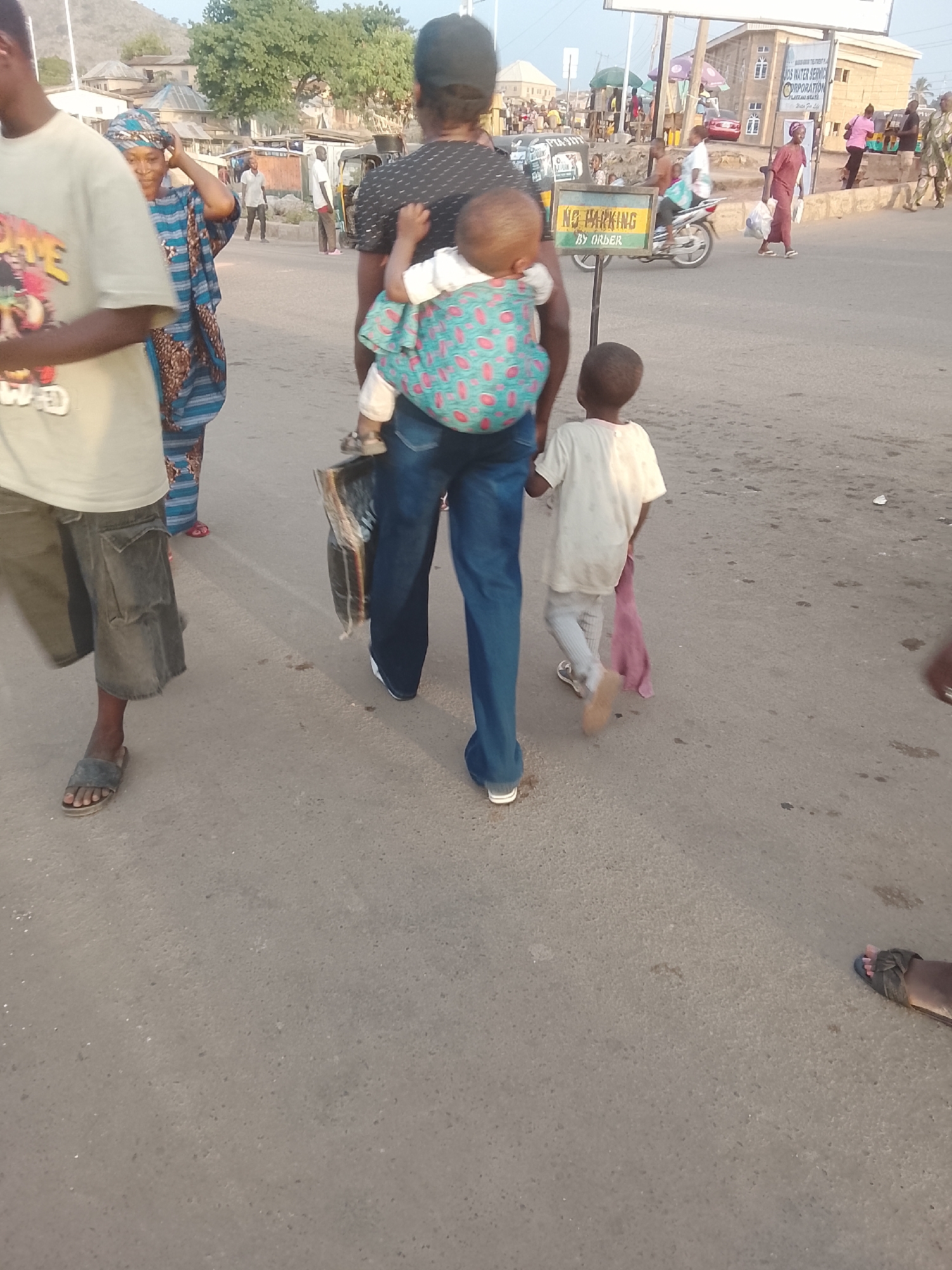 Captured at an eye-level perspective, the image shows an outdoor urban street in Gwong, Nigeria. An individual, facing away from the camera, carries a baby secured on their back with a colorful wrap. A child walks next to the individual, slightly behind. There are other pedestrians present on the street, including one in traditional Nigerian clothing. Objects visible include a "No Parking by Order" sign, a motorcycle, buildings, and a distant hill. The ground is paved and shows signs of wear and dirt. The light suggests it is daytime.