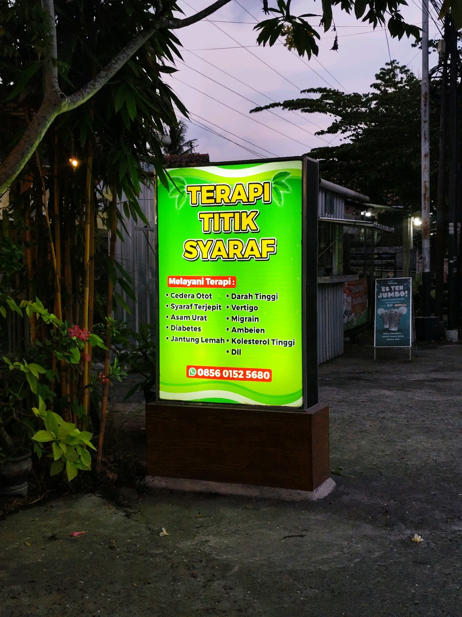 The image shows a glowing green sign that reads "TERAPI TITIK SYARAF" (Nerve Point Therapy) in yellow lettering. The sign lists various conditions treated: "Cedera Otot" (Muscle Injury), "Syaraf Terjepit" (Pinched Nerve), "Asam Urat" (Gout), "Diabetes", "Jantung Lemah" (Weak Heart), "Darah Tinggi" (High Blood Pressure), "Vertigo", "Migrain", "Ambeien" (Hemorrhoids), and "Kolesterol Tinggi" (High Cholesterol), with "Dll" (Etc.) at the end. A phone number, "0856 0152 5680", is also displayed in red. The sign is mounted on a dark brown wooden base.
The sign is situated outdoors at dusk, with the sky showing a gradient of purple and orange. To the left of the sign, there are dark green leafy plants and bamboo stalks. To the right of the sign, a smaller, freestanding white sign advertises "ES TEH JUMBO!" (Jumbo Iced Tea) with an illustration of a cup and a straw. In the background, there are some structures with corrugated metal roofs and a visible pole with wires, suggesting an urban or semi-urban setting. The ground is paved with a rough, dark material, and a few fallen leaves and flower petals are scattered on it. The overall impression is of a small business, likely a clinic or therapy center, operating in the evening.
The location is identified as Bambanglipuro, Indonesia, based on the presence of the sign which likely serves a local community.