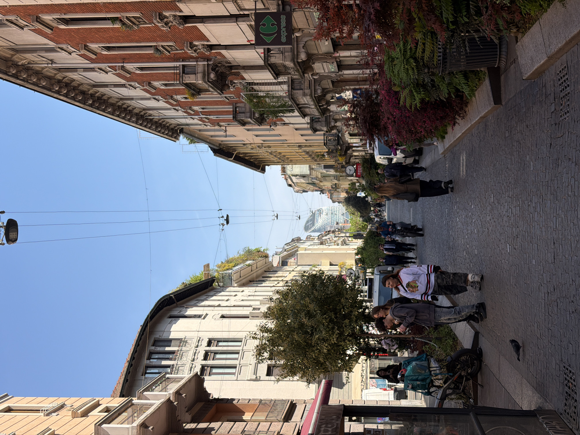 The image shows a street scene in Milan, Italy. The view looks down a narrow street lined with buildings of varying heights and architectural styles. The sky is clear and blue. Overhead wires run the length of the street. People are walking along the cobblestone sidewalk. A man wearing a Chicago Blackhawks jersey stands near a tree and other people. A bicycle laden with items is parked on the sidewalk. A sign for "express" is visible on one of the buildings. In the distance, a modern skyscraper can be seen.