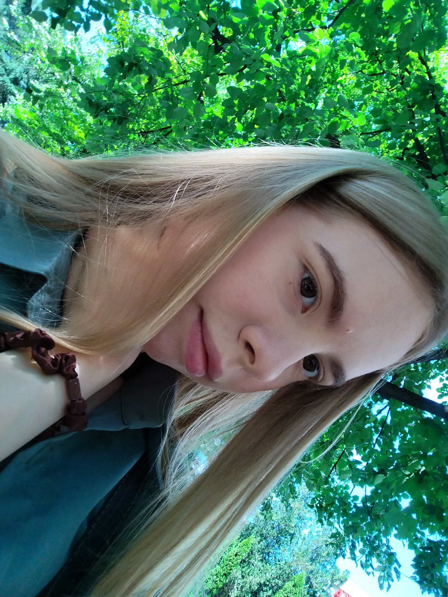 A young person with long blonde hair is looking at the camera, tilted to the side. They are outdoors, with a backdrop of lush green trees and a bright blue sky. The person is wearing a teal shirt and a dark brown scrunchie on their wrist. Their expression is neutral, with their lips slightly parted. The sunlight filters through the leaves, creating dappled patterns on the scene. The time of day appears to be daytime, with bright lighting suggesting fair weather. The overall impression is one of casual serenity, perhaps a moment captured while relaxing in nature. There is no visible text in the image.