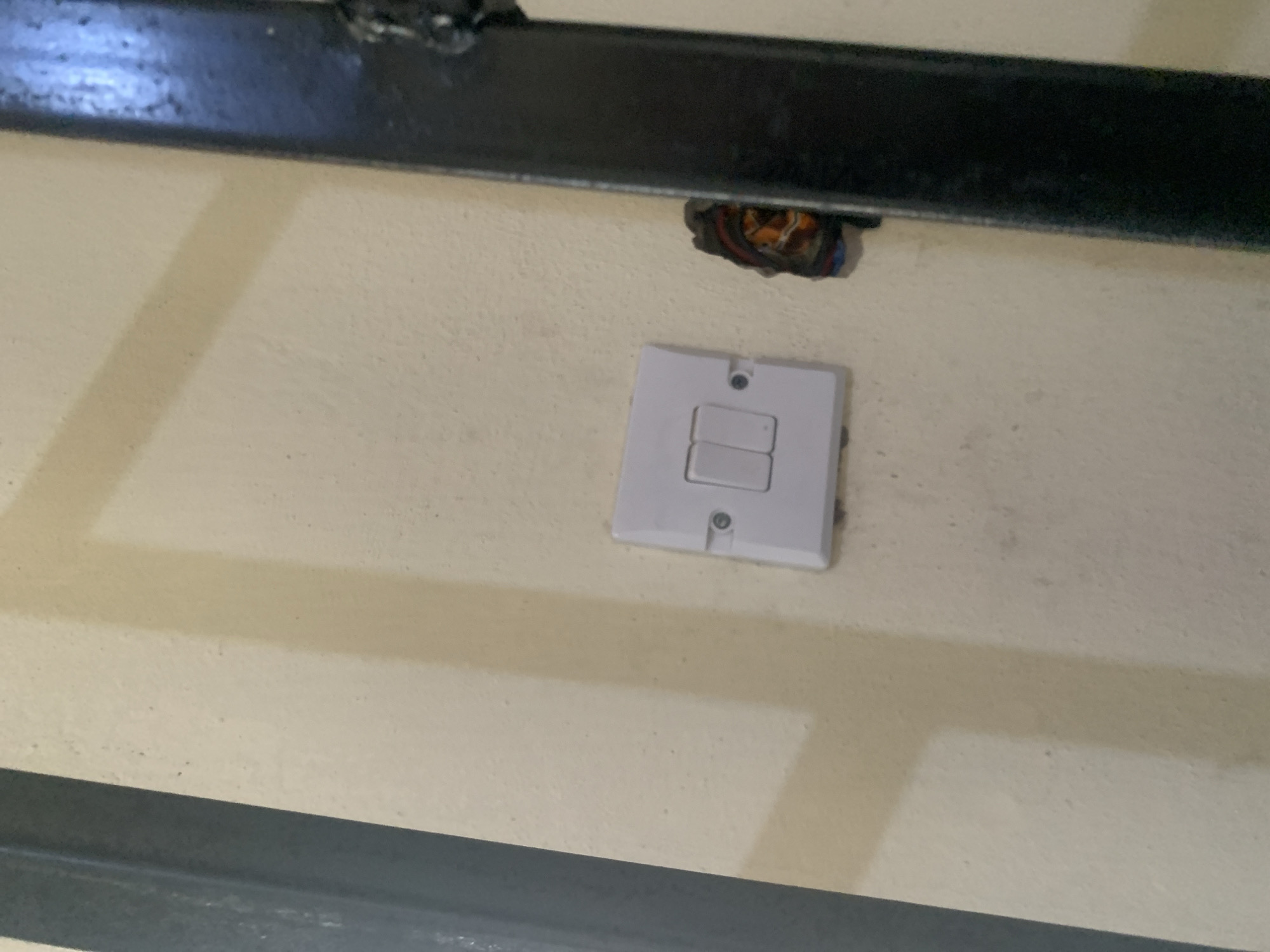 The image displays a close-up of a wall with a light switch. The wall is painted a light cream color and shows some texture. To the left of the image, a dark gray metal bar runs vertically. To the right of the image, another dark metal bar is visible, also running vertically, and appears to be part of a structure or furniture. Adjacent to the light switch, on the right side, there is an exposed bundle of electrical wires. The light switch is a standard white, dual-rocker switch. The location is Jauro Yinu, Nigeria.