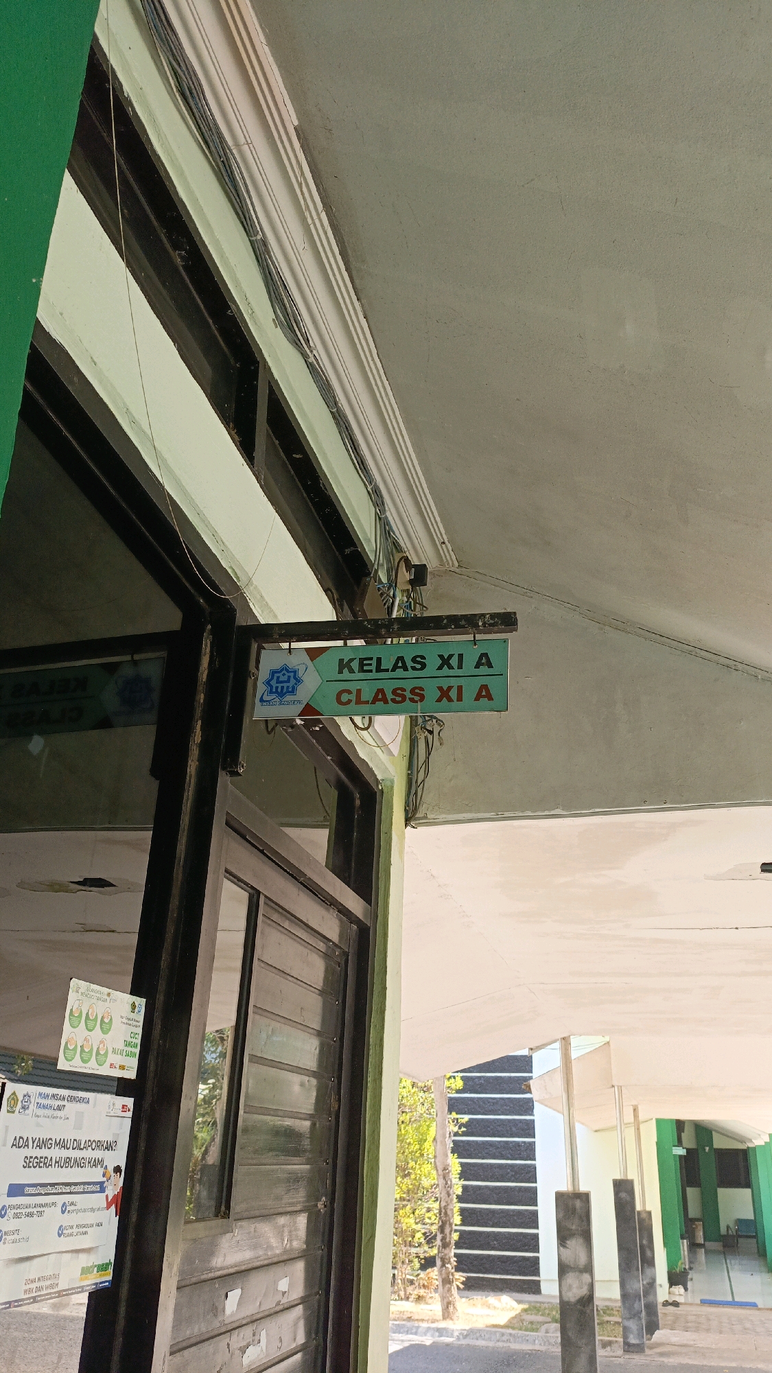 The image shows a building exterior with a sign that reads "KELAS XI A CLASS XI A" in Indonesian and English, indicating a classroom designation. The sign is green with white text and a blue and white logo. The building has a light green painted facade and a dark-framed glass door. Wires are visible running along the ceiling. To the left of the door, there are several posters attached to the wall. One poster has an illustration of babies with the text "LANGKAH-LANGKAH PP..." and "CICI TANGAN PAKAI SABUN". Another poster, partially obscured, contains text including "MAN INSAN CENDEKIA TANAH LAUT", "ADA YANG MAU DILAPORKAN?", and contact information. The background shows an outdoor walkway with pillars and a building with a dark, textured wall. The environment appears to be sunny. The location is identified as Desa Pabahanan, Pelaihari, Tanah Laut, Indonesia. There are no people visible in the image.