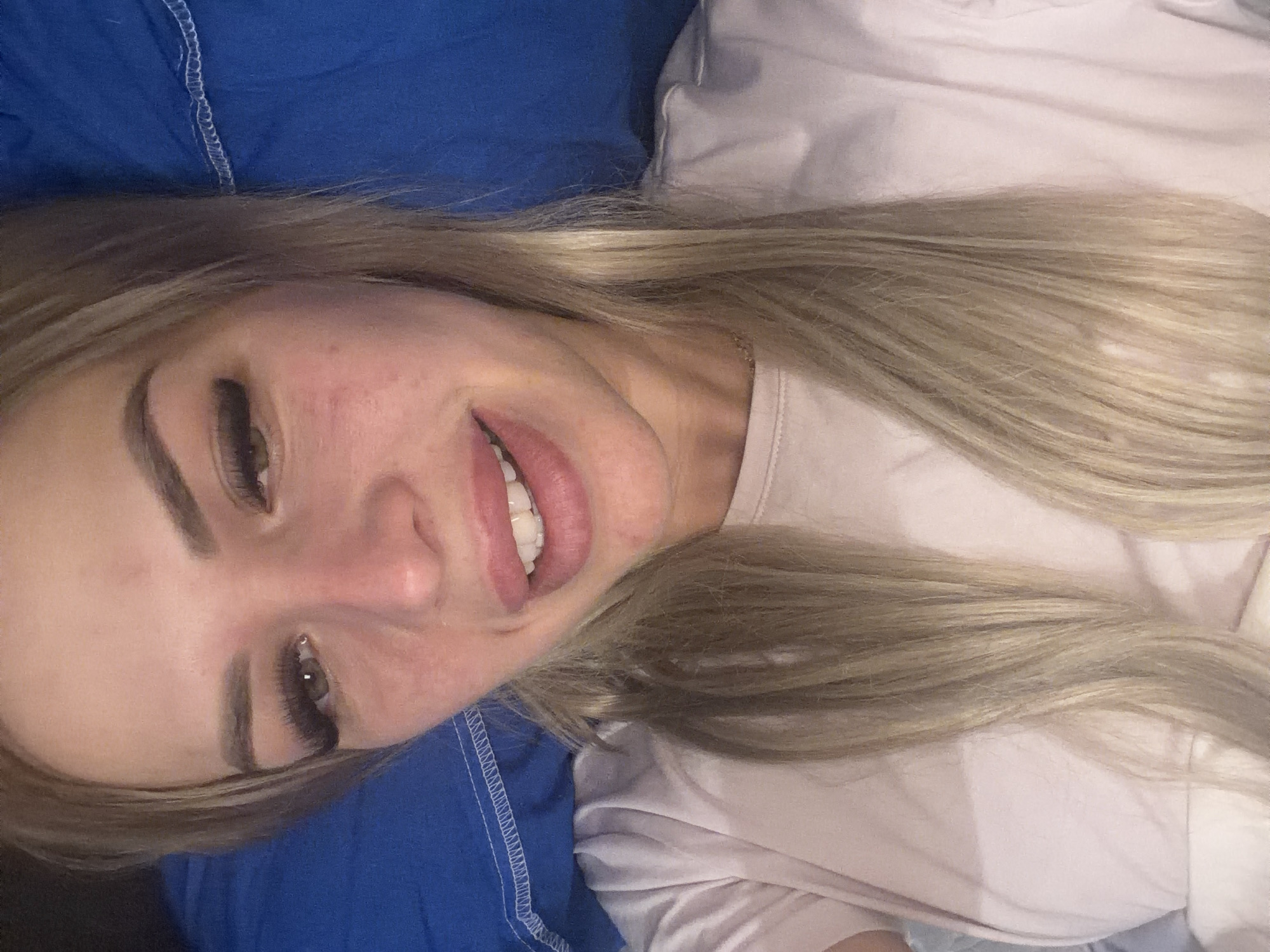 The image shows a close-up, overhead shot of a woman with blonde hair, lying down with her head on a blue pillow. She is wearing a light pink t-shirt. Her eyes are open, and she is looking towards the camera with a slight smile. Her makeup includes visible eyelashes and lip gloss. The background consists of the blue pillow and the edge of what appears to be a white sheet or blanket. The lighting suggests an indoor setting, possibly a bedroom. There are no identifiable landmarks or contextual clues to place the image in Kyiv, Ukraine, or any specific city.