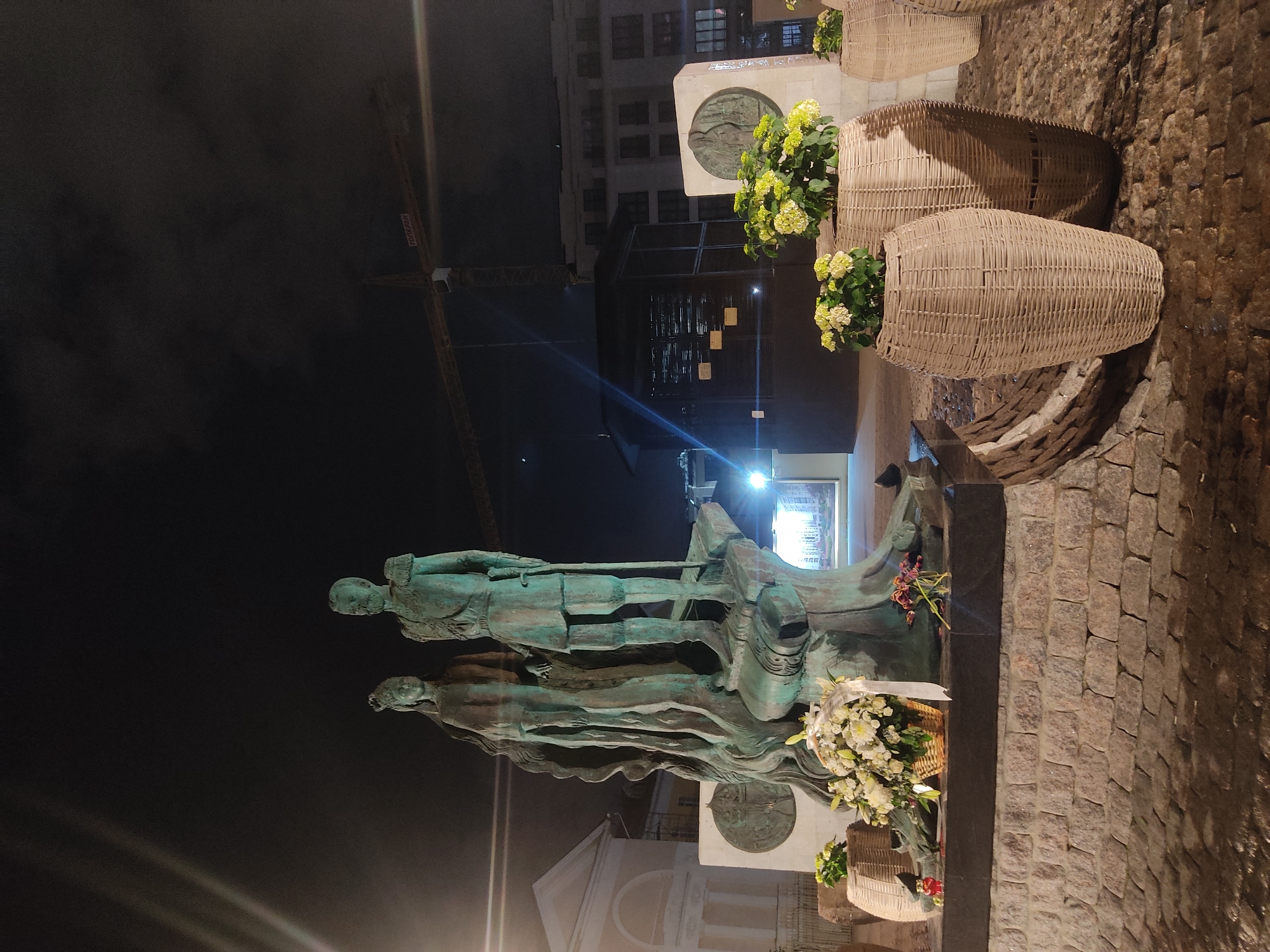 The image depicts a bronze statue of two figures at night in Moscow, Russia. The statue is illuminated by artificial lights and is set against a dark background. In the background, there are buildings and a construction crane. The statue is surrounded by floral arrangements in woven baskets, some of which are overflowing. The ground is paved with cobblestones. The statue is also on top of a stone structure with a circular plaque on the front.