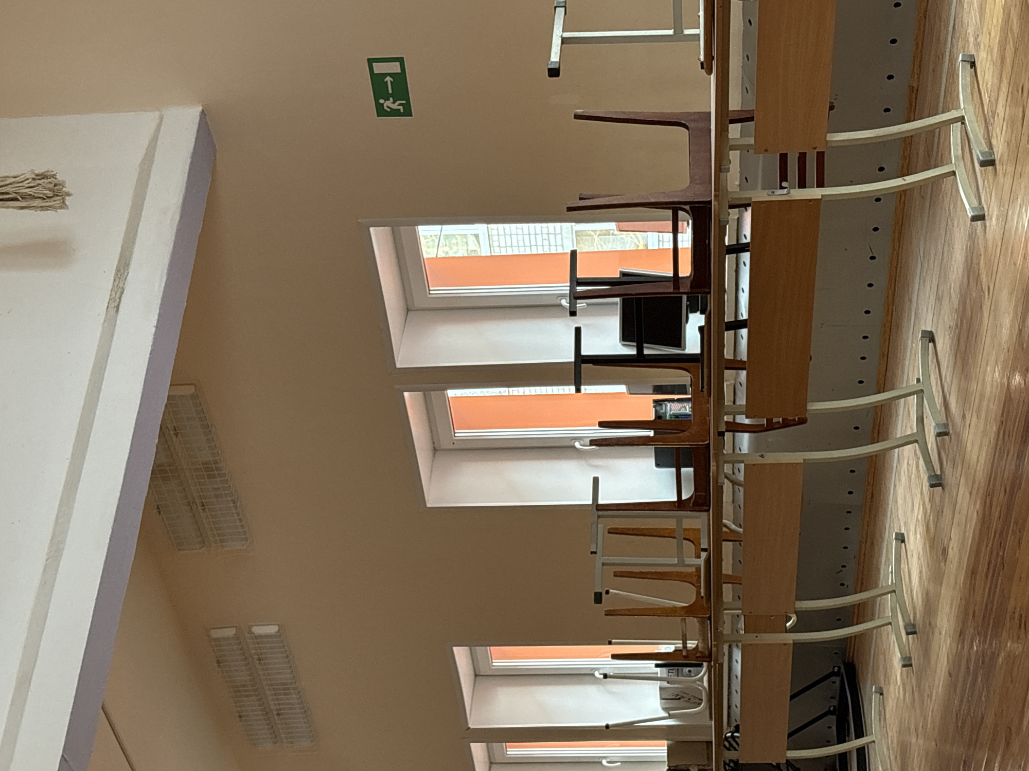 The image depicts the interior of a room, possibly in a school or community center in Chaikovsky, Russia. The room has a row of skylight windows with orange tints, stacked chairs and desks, and fluorescent ceiling lights. The chairs are stacked on top of the desks, likely for storage or cleaning purposes. A green emergency exit sign is visible on the wall. The floor is made of wood.