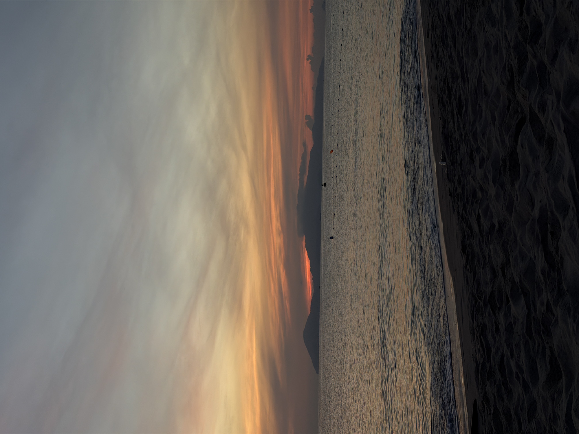 This is a photograph of a beach at sunrise, likely taken in Hoi An, Vietnam. The sky is painted with warm hues of orange and yellow, transitioning to a soft gray at the top. Wispy clouds stretch across the horizon. In the distance, silhouetted mountains rise from the sea, their shapes softened by the morning mist. The water is calm, with gentle waves lapping at the shore. The foreground is a dark, sandy beach, with the texture of the sand clearly visible. There are no people or discernible objects in the immediate vicinity of the foreground or mid-ground. Some buoys or markers are visible in the water, suggesting a designated swimming or boating area. The overall atmosphere is serene and tranquil.