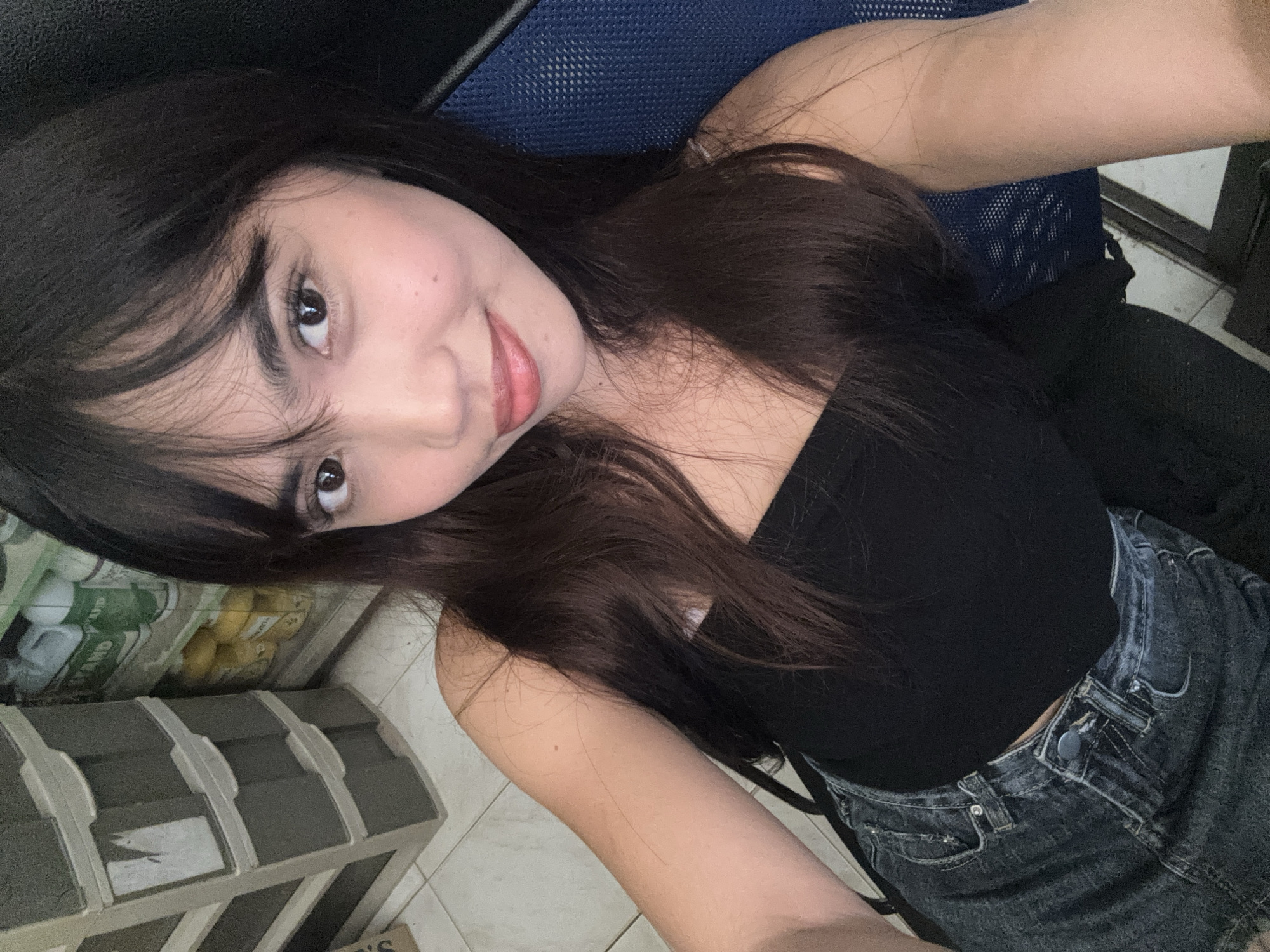 A young woman with long, dark hair and bangs is taking a selfie in an indoor setting. She is wearing a black strapless top and denim shorts. To her left, a gray plastic storage unit with multiple drawers is visible. Behind her, on the right, a portion of a blue mesh chair is present. The flooring appears to be tiled. The background also shows shelves with various items, including bottles and containers, suggesting a storage area or a small shop. The overall lighting is artificial. There are no discernible landmarks indicating the specific location within Tagum City, Philippines.