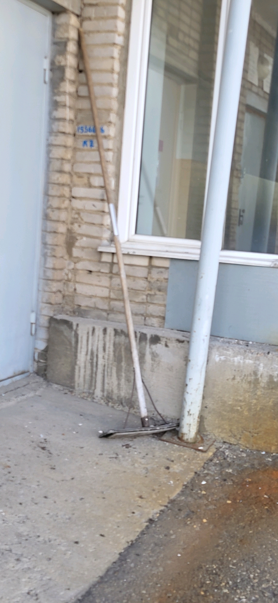 The image depicts an outdoor scene near a building in Oskemen, Kazakhstan. A rake with a metal head and a wooden handle leans against a brick wall and a white pipe. The wall has blue markings that appear to be numbers and letters: "15566" and "KZ". In front of the wall is a concrete ledge with visible water streaks. The ground in the foreground is concrete, with some debris scattered on it. To the right, there is an area of asphalt with a wet appearance, possibly indicating a puddle or recent rain. The building features a white-framed window. The overall impression is a utilitarian exterior space.