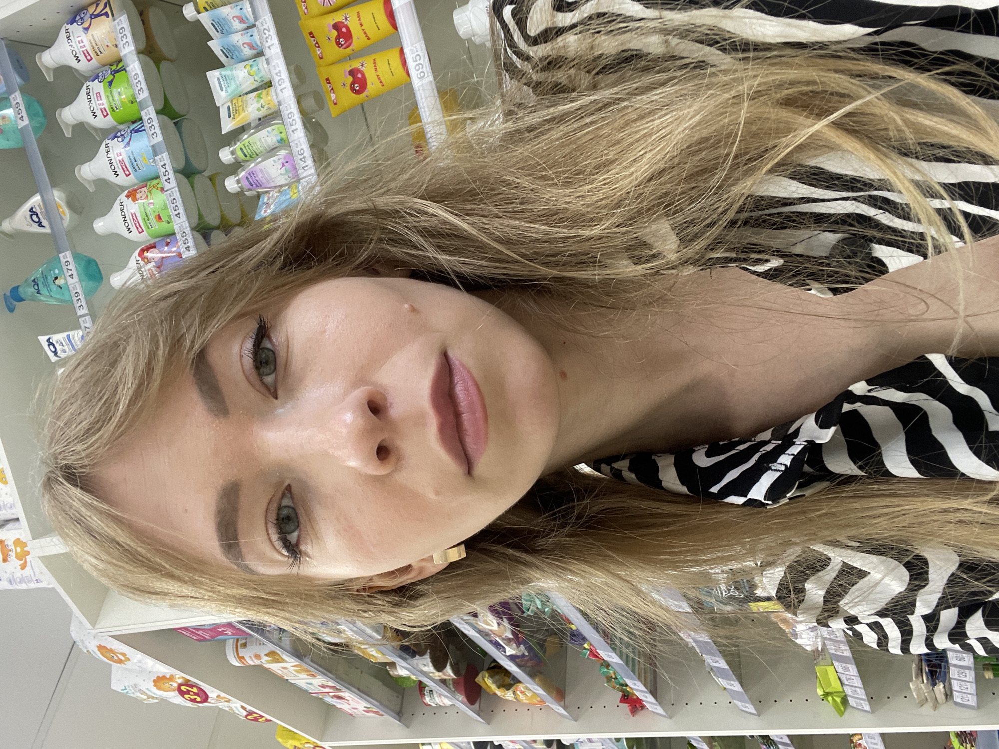 The image shows a young woman with blonde hair and green eyes taking a selfie in a store aisle. She is wearing a black and white zebra-print shirt and a gold earring. The background consists of shelves stocked with various products, including bottles of shampoo and tubes of cream. The overall scene suggests a retail environment, possibly a pharmacy or a supermarket. The lighting is bright and even, indicating indoor lighting. There are no other people visible in the frame. The woman's facial expression is neutral, and she is looking directly at the camera. The image is taken from a slightly low angle, with the woman's face filling a significant portion of the frame.