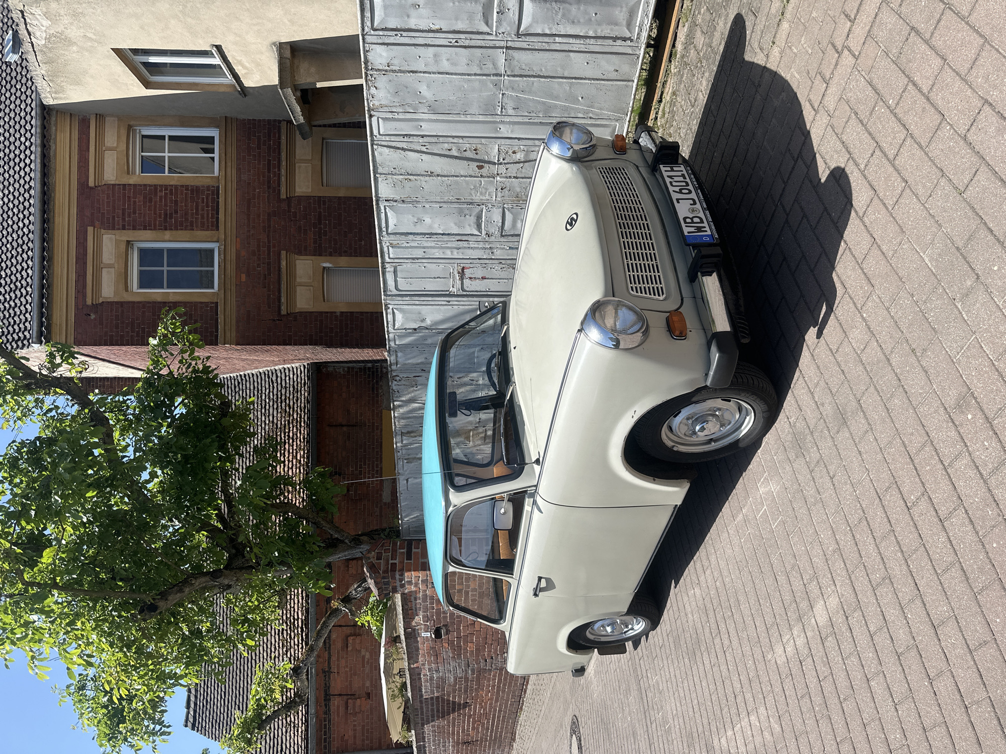 A light-colored Trabant car with a blue roof is parked on a brick-paved surface in Jessen (Elster), Germany. The car faces to the right in the frame. Buildings of brick and plaster construction are visible in the background, along with a large tree with green leaves. The street is lined with sidewalks. The license plate of the car is partially visible, reading "WB J 601H." A shadow cast by the car falls onto the paved surface. The weather appears sunny.
