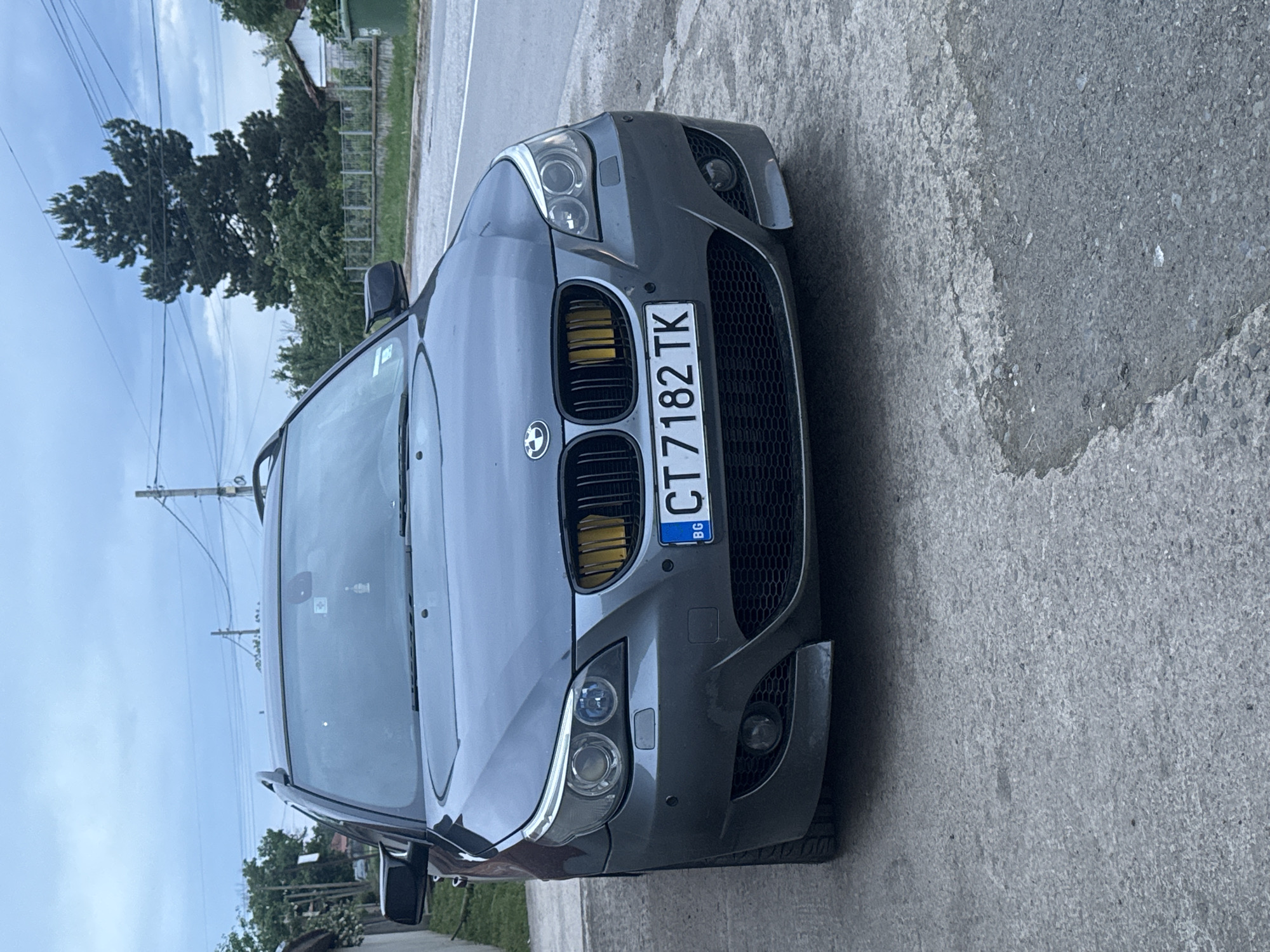 The image shows a gray BMW with a Bulgarian license plate "CT 7182 TK". The car is parked on a concrete surface next to a road. The background includes trees, a building, and power lines against a cloudy sky. The image suggests an outdoor setting, likely in an urban area. The car is located in the city of Sarnevo, Bulgaria.