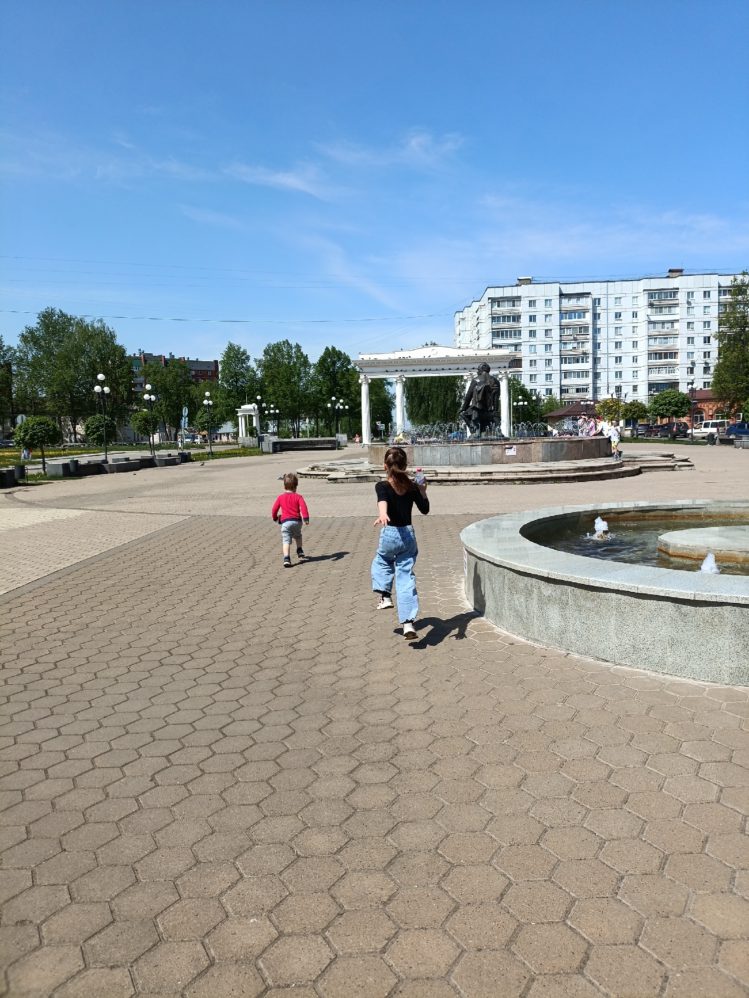 The image depicts a sunny outdoor scene in what appears to be a public square in the city of Chaikovsky, Russia. The ground is paved with hexagonal stone tiles. Two people are present: a child in a red shirt and grey pants runs ahead of a taller individual, possibly a young adult, wearing a black shirt and light blue jeans. Both are moving away from the viewer towards a large fountain with a statue in the center. The fountain is surrounded by a white, colonnaded structure. Another smaller, circular fountain is visible in the foreground on the right. Behind the main fountain, there are trees and a large, multi-story apartment building. Several other people are gathered near the fountain area. Lampposts are placed intermittently around the square. The sky is blue with some clouds. The scene appears to capture a candid moment of children playing in an urban environment.