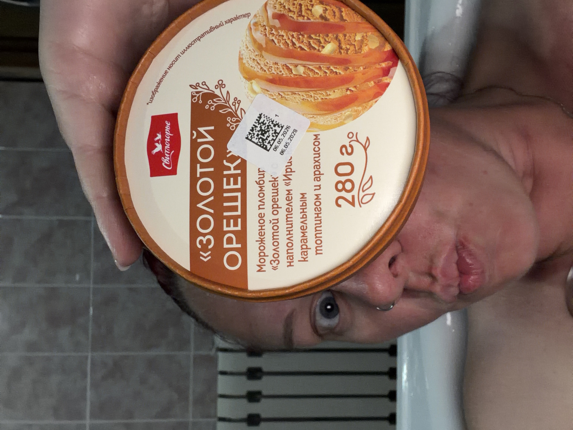 A person is holding a container of "Golden Nut" ice cream (280g) made by "Svitlogorie" brand. The container has an image of the ice cream and a sticker with expiration dates: 06.05.2026 and 06.05.2028. The person's face, partially obscured by the ice cream container, shows their left eye and mouth. The background consists of a tiled wall and what appears to be a part of a white bathtub. The image was likely taken in a bathroom. No definitive information is present in the image to confirm its location in the city of Chaikovsky, Russia.