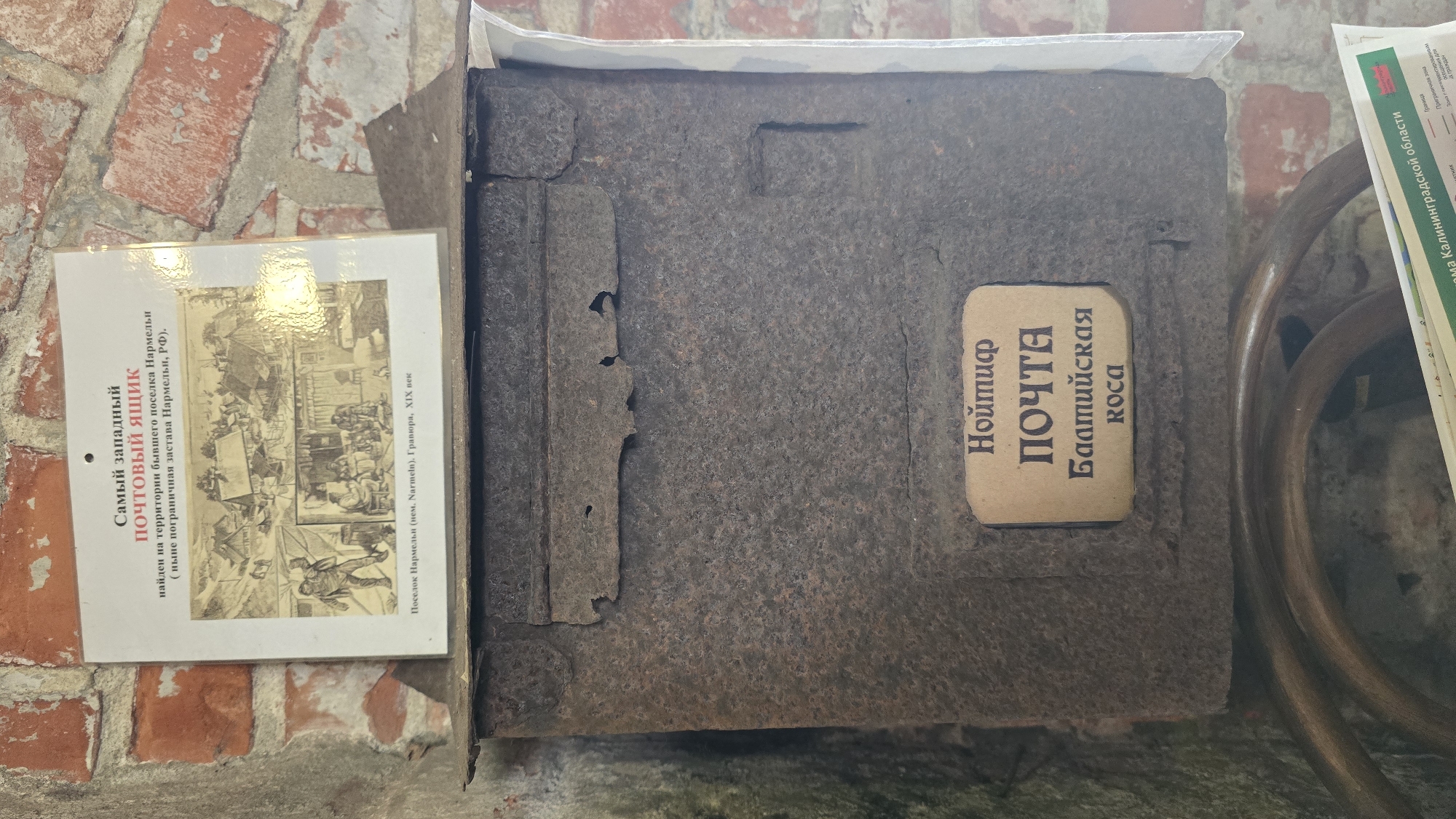 An old, heavily rusted metal post box is the central subject, displayed against a backdrop of a reddish-brown brick wall. To its left, a white informational sign provides historical context. On the right, parts of a dark, curved wooden or metal structure and a green-and-white map are visible.
The post box features a pale rectangular label with Cyrillic text: "Нойтиф" (Neuhäuser), "ПОЧТА" (POST), "Балтийская" (Baltic), and "KOCA" (Spit), suggesting it is a historical post box from Neuhäuser on the Baltic Spit.
The informational sign is titled "Самый западный ПОЧТОВЫЙ ЯЩИК" (The westernmost MAILBOX). The text explains it was "найден на территории бывшего поселка Нармельн (ныне пограничная застава Нармельн, РФ)" (found on the territory of the former village Narmeln, now a border outpost in Narmeln, RF). Below this description are several small, black-and-white engravings, identified as depicting "Поселок Нармельн (нем. Narmeln). Гравюра, ХІХ век" (Narmeln village (Ger. Narmeln). Engraving, XIX century).
A fragment of a map or document on the right includes phrases like "Дома Калининградской области" (Houses of Kaliningrad Oblast) and "Приграничная зона" (Border zone), further reinforcing the location context as the Kaliningrad region of Russia, specifically near Baltiysk. The scene depicts a historical exhibit, likely in a museum, focusing on a significant postal artifact from the region.
