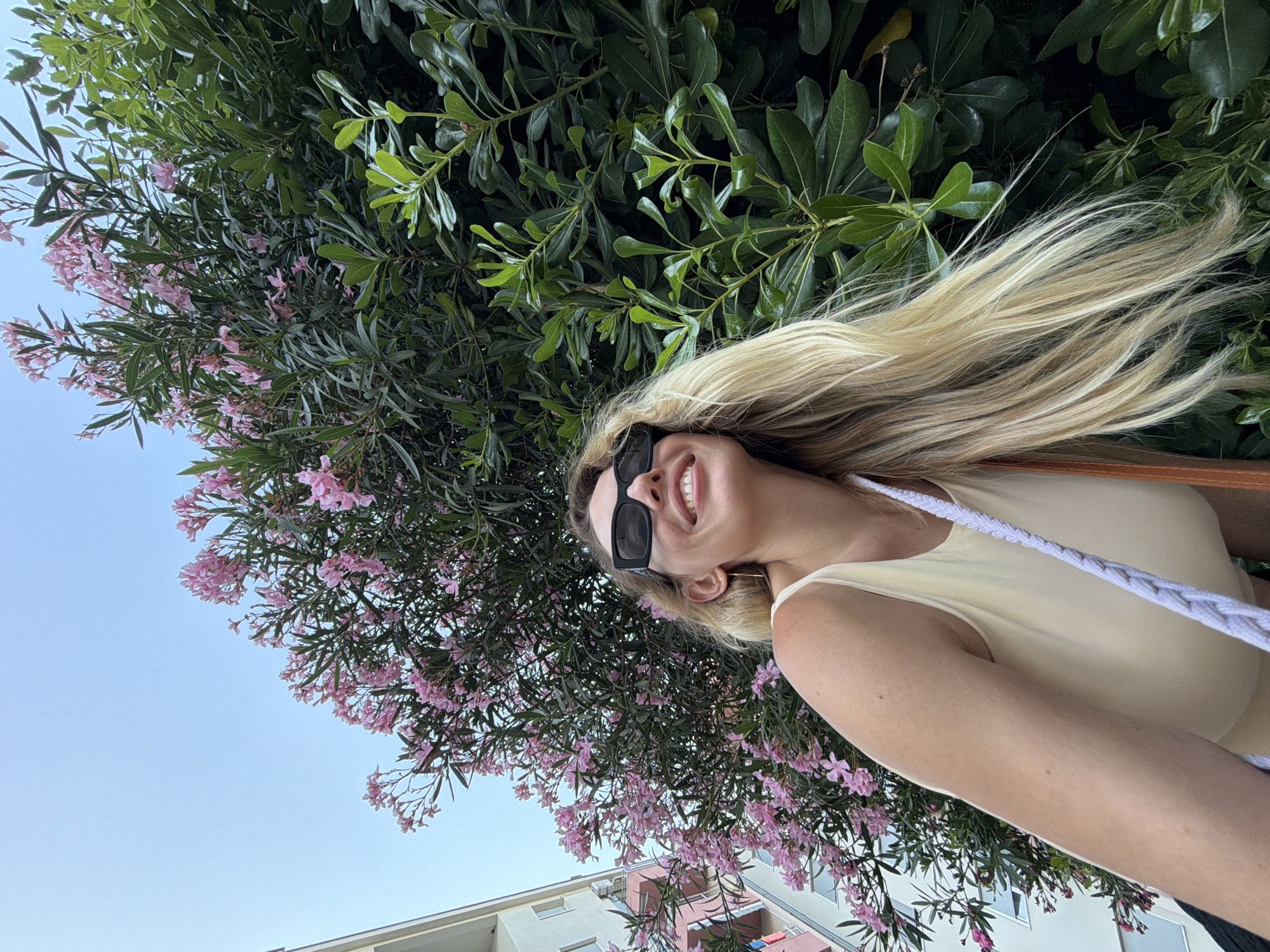 In a low-angle, upward shot, a blonde woman with sunglasses smiles joyfully, looking up towards a pink blooming oleander bush. She wears a light-colored tank top and a purse strap is visible over her shoulder. Behind her, the facade of a pale pink and white building is partially visible. The scene appears to be in Budva, Montenegro, suggested by the architecture and lush greenery commonly found in the region. The sky above is a clear, pale blue.
