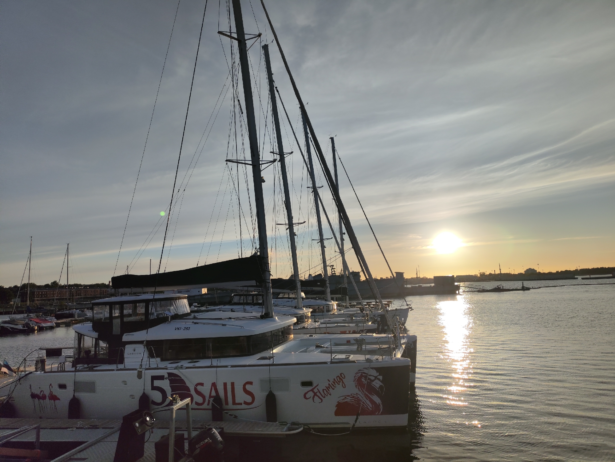 A fleet of white catamarans with tall masts are docked in a harbor, positioned parallel to the shore and facing right. The boats are predominantly white with dark-tinted windows and prominent red branding, including the number "5" and the word "SAILS" on the side of the nearest vessel. A stylized flamingo graphic adorns the stern of this catamaran, along with the word "Flamingo". The sun is setting in the background, casting a warm glow and creating a shimmering reflection on the calm water. The sky is partly cloudy with wispy cirrus clouds. In the distance, a city skyline with industrial elements and cranes is visible. A small boat with a red cover is docked to the left. The overall scene depicts a tranquil evening in a marina.