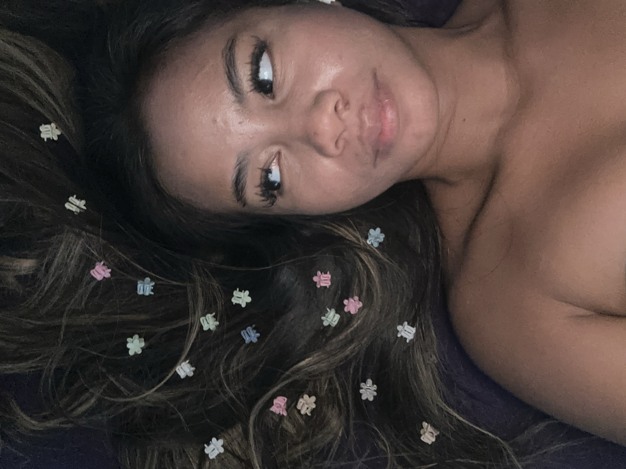 A woman lies on a purple surface. Her long, dark hair is adorned with colorful, small, plastic hair clips. The image focuses on her face, which is angled towards the camera. Her eyes look to the right. The background is out of focus. There is no verifiable information to suggest this image originated in North Jakarta, Indonesia. The image provides no location context.