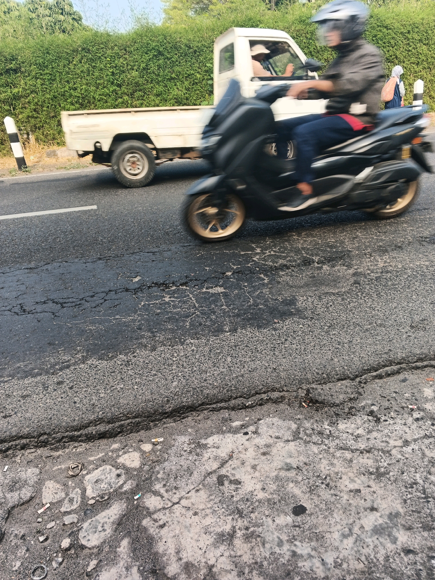 The image captures a street scene in Ungaran, Indonesia, characterized by a damaged asphalt road surface. In the foreground, the road is visibly cracked and uneven. A white pickup truck is partially visible on the left side of the frame, its rear section and wheel showing. To the right, a person on a black motorcycle is in motion, their body and the vehicle blurred due to the movement. The rider is wearing a helmet and dark clothing. In the background, a dense green hedge forms a backdrop, and a person with a backpack is walking away from the motorcycle. A white pole with black markings stands near the hedge on the left. The sky is overcast and the overall lighting suggests it is daytime.