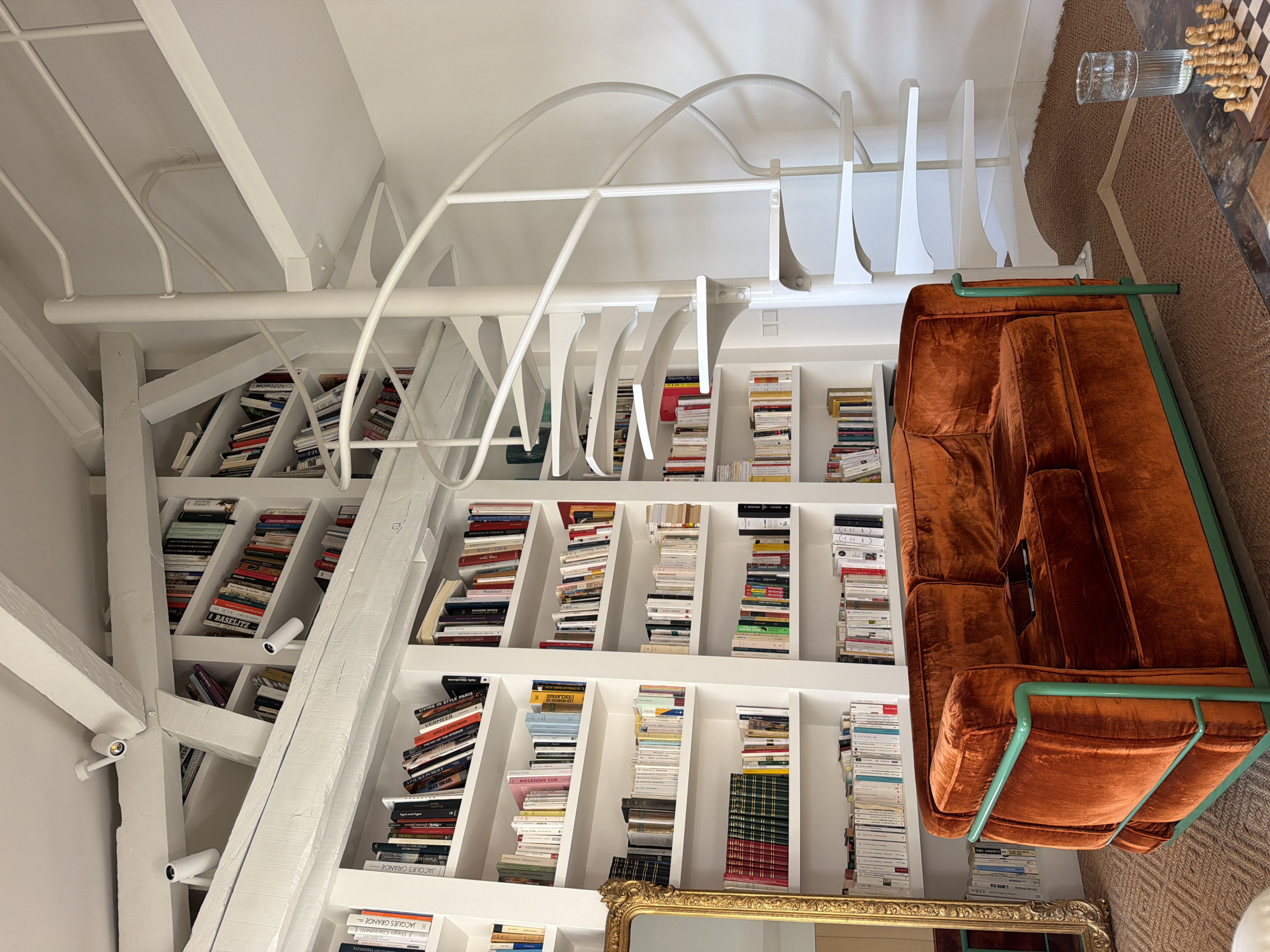 Here is a forensic description of the image:
The interior shot depicts a room with a large, white, built-in bookshelf filled with books. The books are arranged in a variety of ways, some vertically and some horizontally, and the shelves have varying depths and angles. A white, structural, curved element with vertical supports spans across the bookshelf, adding an architectural detail. The room appears to have exposed beams and track lighting, also in white.
In the foreground, a vintage-style orange velvet couch with a green frame is partially visible. On the top right of the image, a table with a chessboard, pieces, and a glass of water are present. The floor is covered with a brown textured rug. The bottom center shows a portion of a gold-framed mirror.
Books within the bookshelf include titles such as "Diego Giacometti 1" and "JACQUES GRANGE", other titles may be present but are illegible.