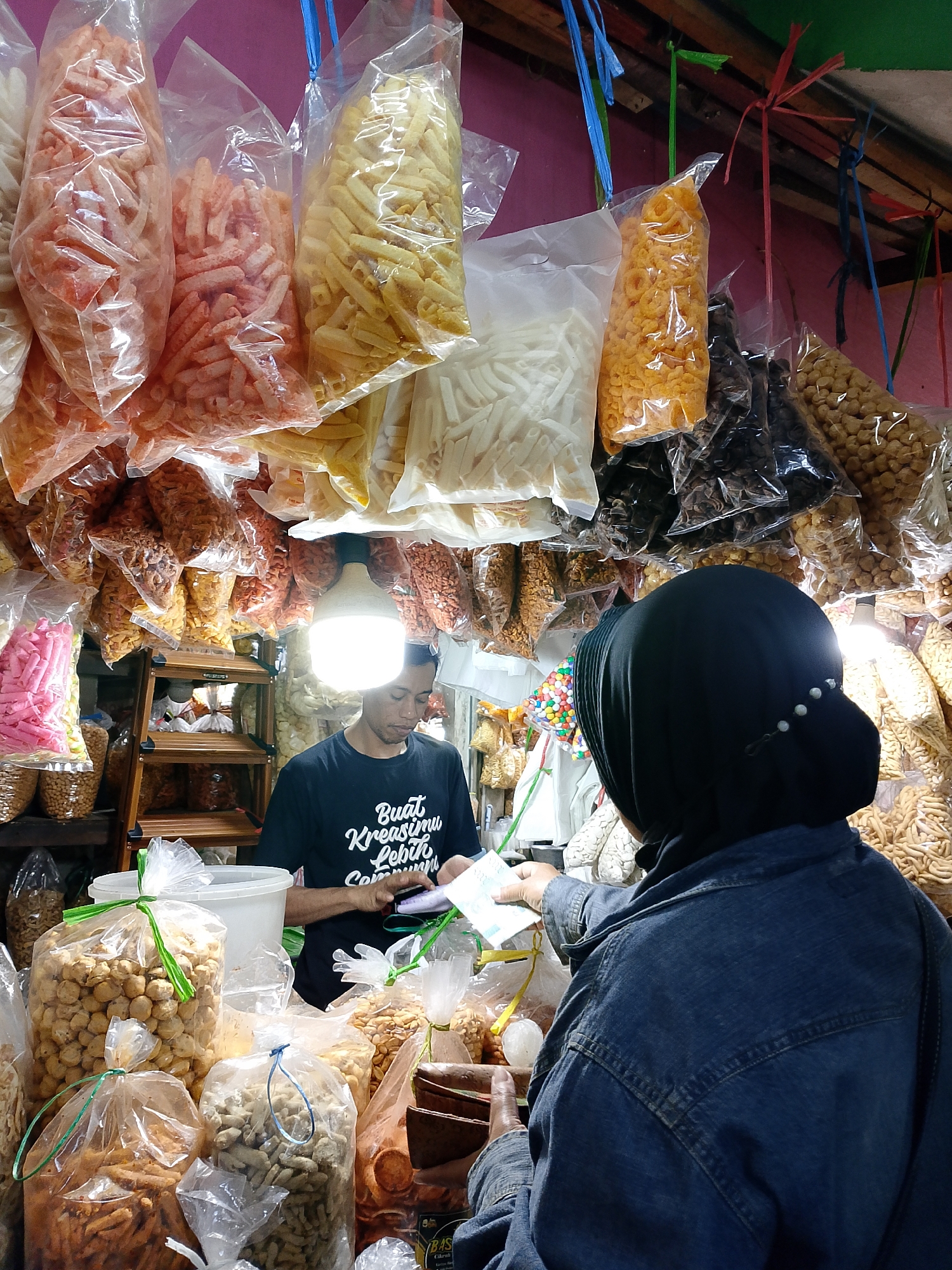 A person wearing a black hijab and denim jacket is exchanging money with a vendor in a street market stall in Surabaya, Indonesia. The stall is filled with bags of snacks, including chips, crackers, and candies, hanging from the ceiling and stacked on shelves. A bright light bulb illuminates the scene. The vendor, a man wearing a black t-shirt, is holding out a small stack of bills and a receipt. The customer is reaching out to take them.