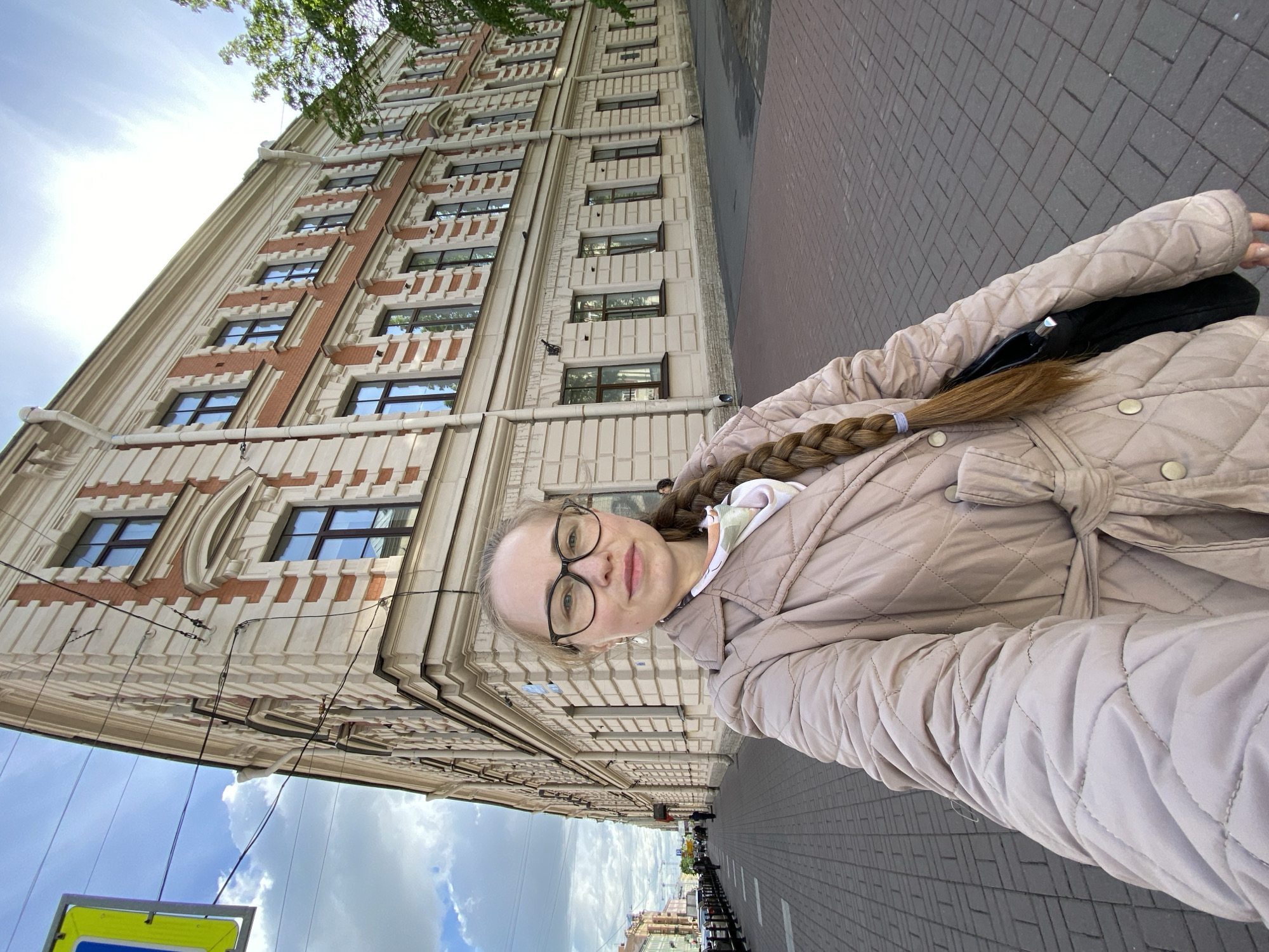 A medium shot captures a woman wearing glasses and a light beige quilted coat standing on a red brick sidewalk in Saint Petersburg, Russia. She is positioned in front of a tall building with multiple windows and decorative architectural details in beige and red. A sidewalk runs along the side of the building, leading into the distance. The sky is partially visible with blue patches and white clouds. Street signs are visible in the lower part of the image. The perspective is tilted, giving the impression that the woman is holding the camera at an angle.