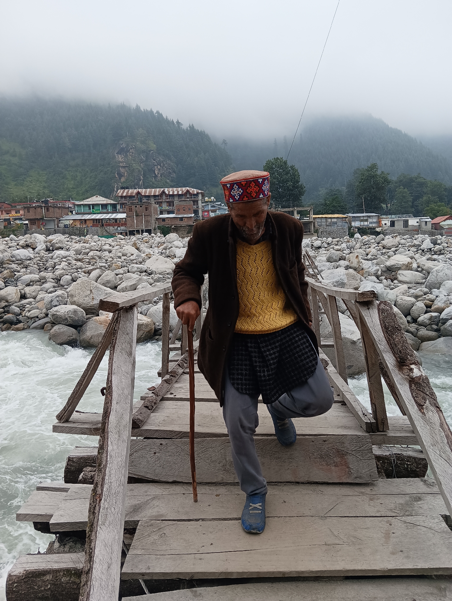 An elderly man with a cane is walking across a wooden bridge in Bahang, India. He is wearing a traditional hat, a yellow sweater, a plaid skirt, and grey pants. The bridge spans a river with white, churning water. The background shows a rocky riverbed and a village with buildings nestled against a misty, forested mountain. The sky is overcast.