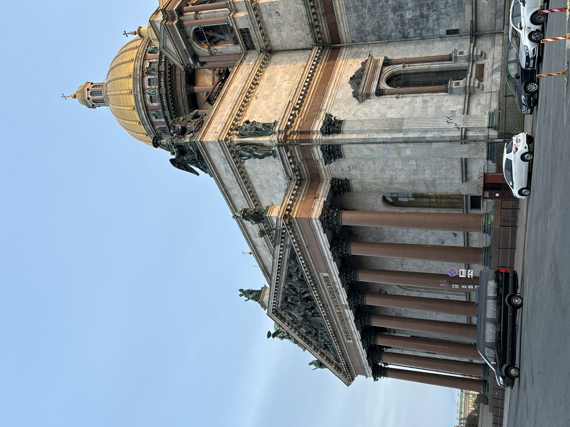 The image shows a partial view of St. Isaac's Cathedral in Saint Petersburg, Russia. The architecture is notable for its large golden dome, classical façade with granite columns, and intricate sculptural details. Several cars are parked along the street adjacent to the cathedral. The sky is light blue.