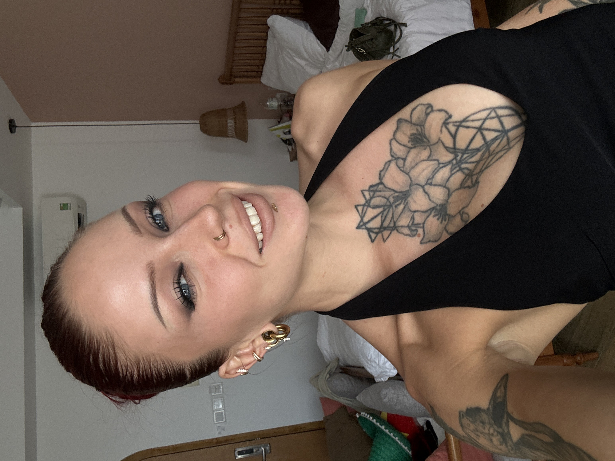 The image shows a close-up selfie of a woman with dark red hair pulled back. She is wearing a black halter top and has several tattoos, including a large floral design on her chest and another on her left arm. She is also wearing multiple earrings and a nose ring, and has lip piercings. The woman is smiling with her mouth slightly open, showing her teeth, and her tongue is sticking out slightly. She is in a room with a bed in the background, a lamp hanging from the ceiling, and what appears to be an air conditioning unit on the wall. The overall setting suggests a hotel room or bedroom. The presence of an air conditioning unit with Vietnamese labeling indicates the photo was likely taken in Vietnam. Given the subject's proximity to the ocean and the common tourist destinations in the region, it's plausible this photo was taken in Da Nang, Vietnam.