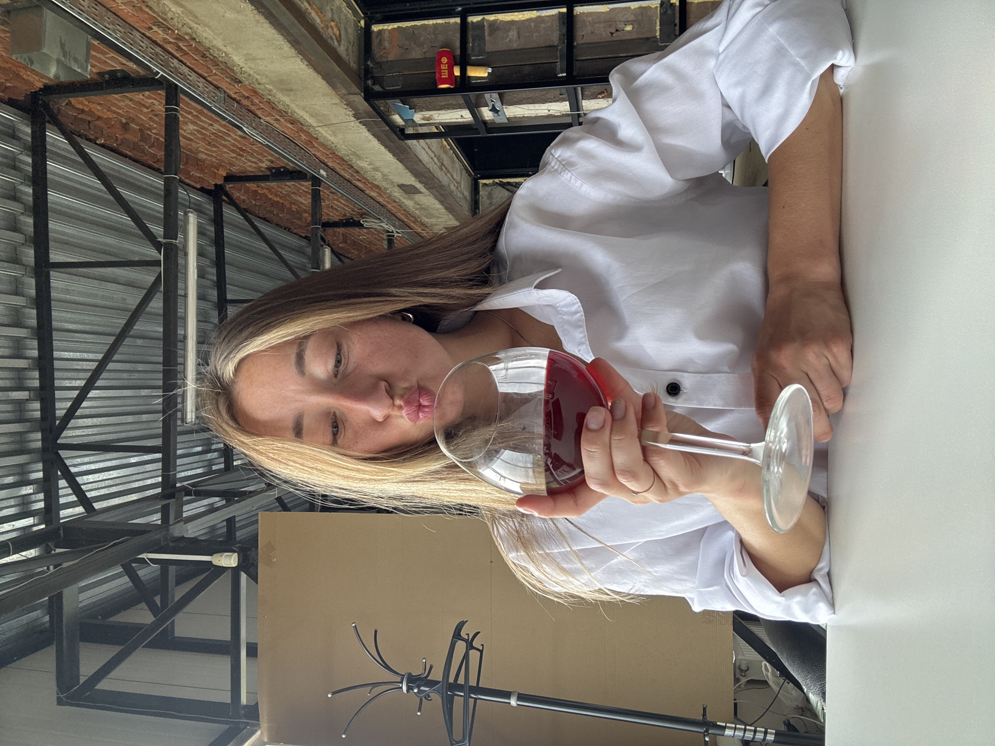 A young woman with blonde hair is holding a glass of red wine in front of her face, with her lips puckered as if to kiss the glass. She is wearing a white collared shirt. The background appears to be a cafe or restaurant with industrial-style exposed beams, corrugated metal ceiling, and brick walls. There is a coat rack to her left and a sign that reads "ШЕФ" (Chef in Russian) to her right. The image was taken in Chaikovsky, Russia.