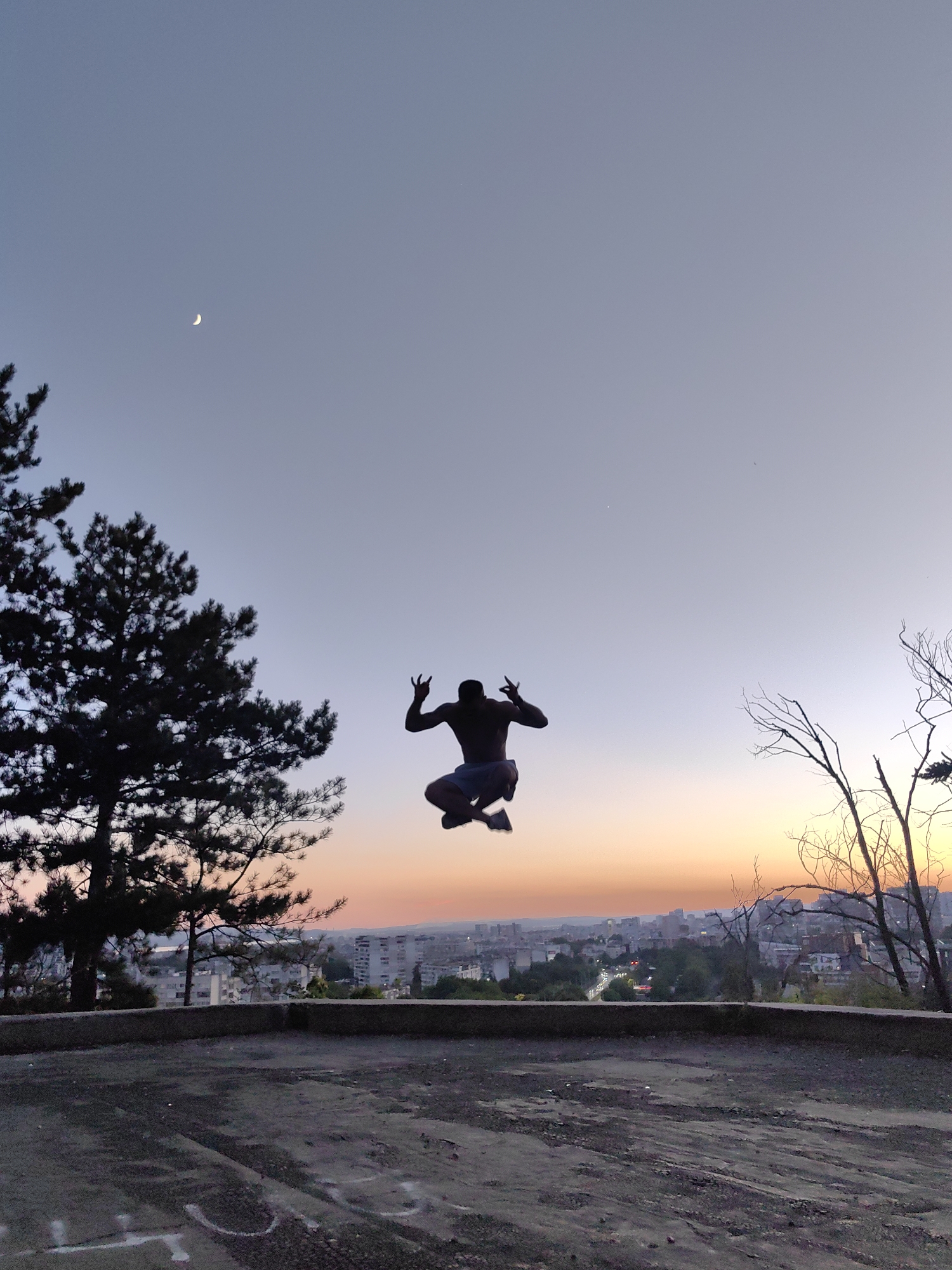 A silhouette of a person is captured mid-air, jumping against a twilight sky above the city of Varna, Bulgaria. The individual, with arms raised in a gesture, is positioned centrally in the frame, with the city's buildings and a faint crescent moon visible in the distance. The foreground shows a concrete surface, possibly a rooftop or elevated platform, with trees on the left and bare branches on the right framing the cityscape. The sky transitions from a pale blue at the top to warm orange and pink hues near the horizon, indicating sunset or sunrise.