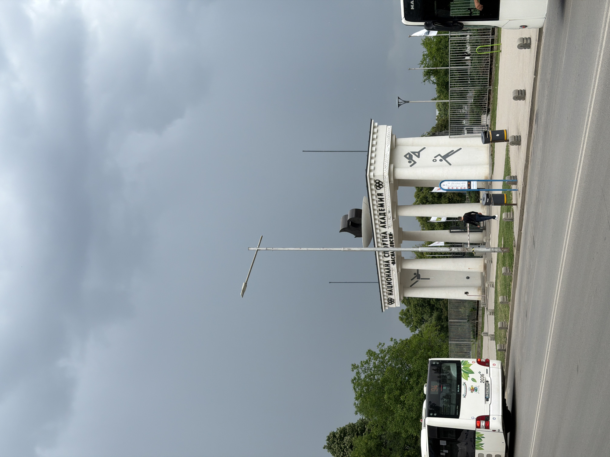 The image shows the entrance to the National Sports Academy in Sofia, Bulgaria. The entrance consists of a white colonnade structure with the Olympic rings and silhouettes of athletes. The name of the academy is written in Bulgarian. A person is walking through the entrance. A white bus is parked on the road next to the entrance. The sky is cloudy and overcast. Trees and foliage are visible in the background. A sidewalk runs along the side of the road, separated by a metal fence. A streetlight stands next to the colonnade structure. A trash can sits on the sidewalk.