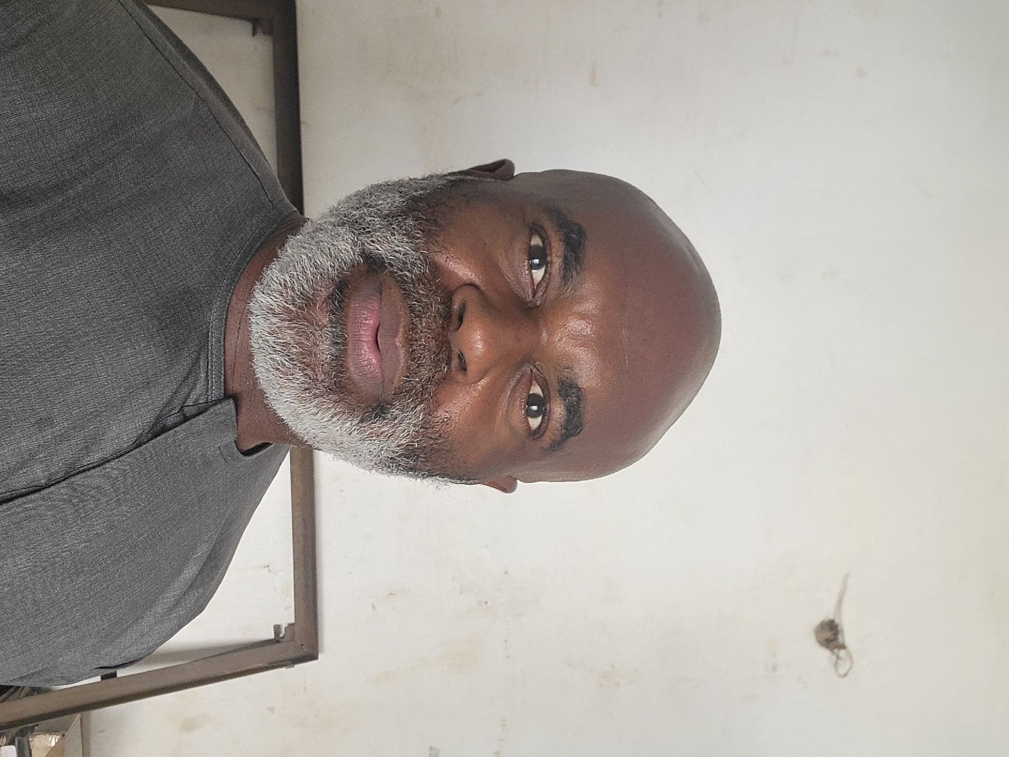 A medium shot captures a dark-skinned man with a shaved head and a full, mostly gray beard. He is wearing a gray shirt. The background is a plain, off-white wall with some smudges and a small object hanging on it. A metal frame is visible on the left edge of the frame. There is no information about the location in the city of Maiburiji, Nigeria in the image.