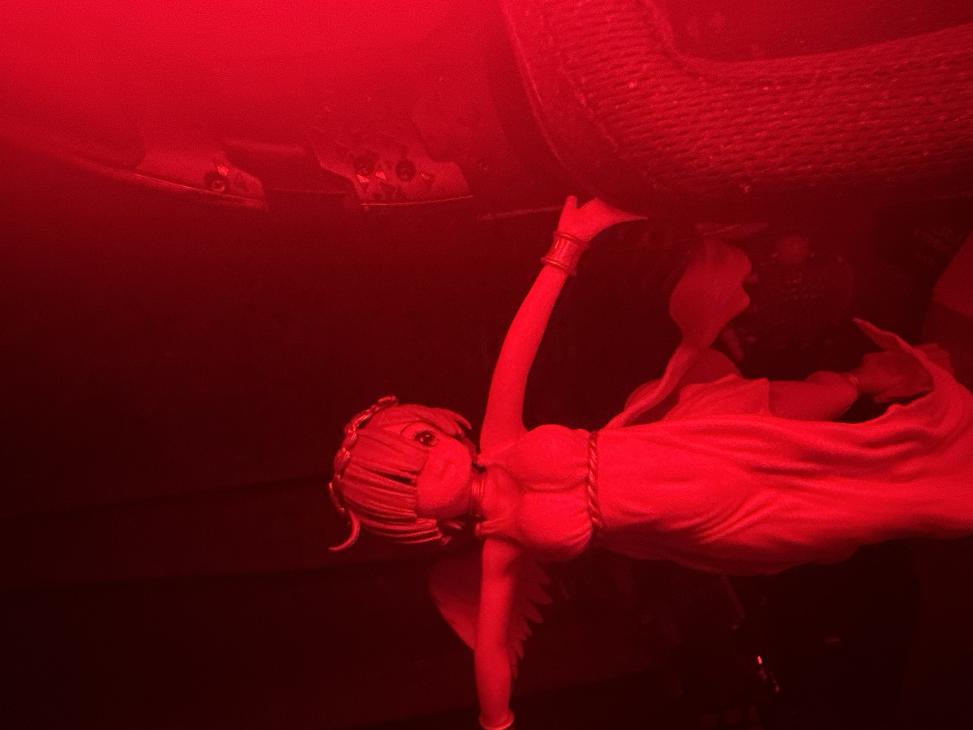 A close-up, low-angle shot shows an angelic figurine illuminated by red light against a dark background. The figurine, with outstretched arms and small wings, wears a white, flowing dress and a band around its head. A mask covers its mouth and nose. To the right of the figurine, a dark, textured object, possibly a rough fabric or rope, occupies the foreground. The overall scene is bathed in a strong red hue, obscuring finer details. The location is unknown.