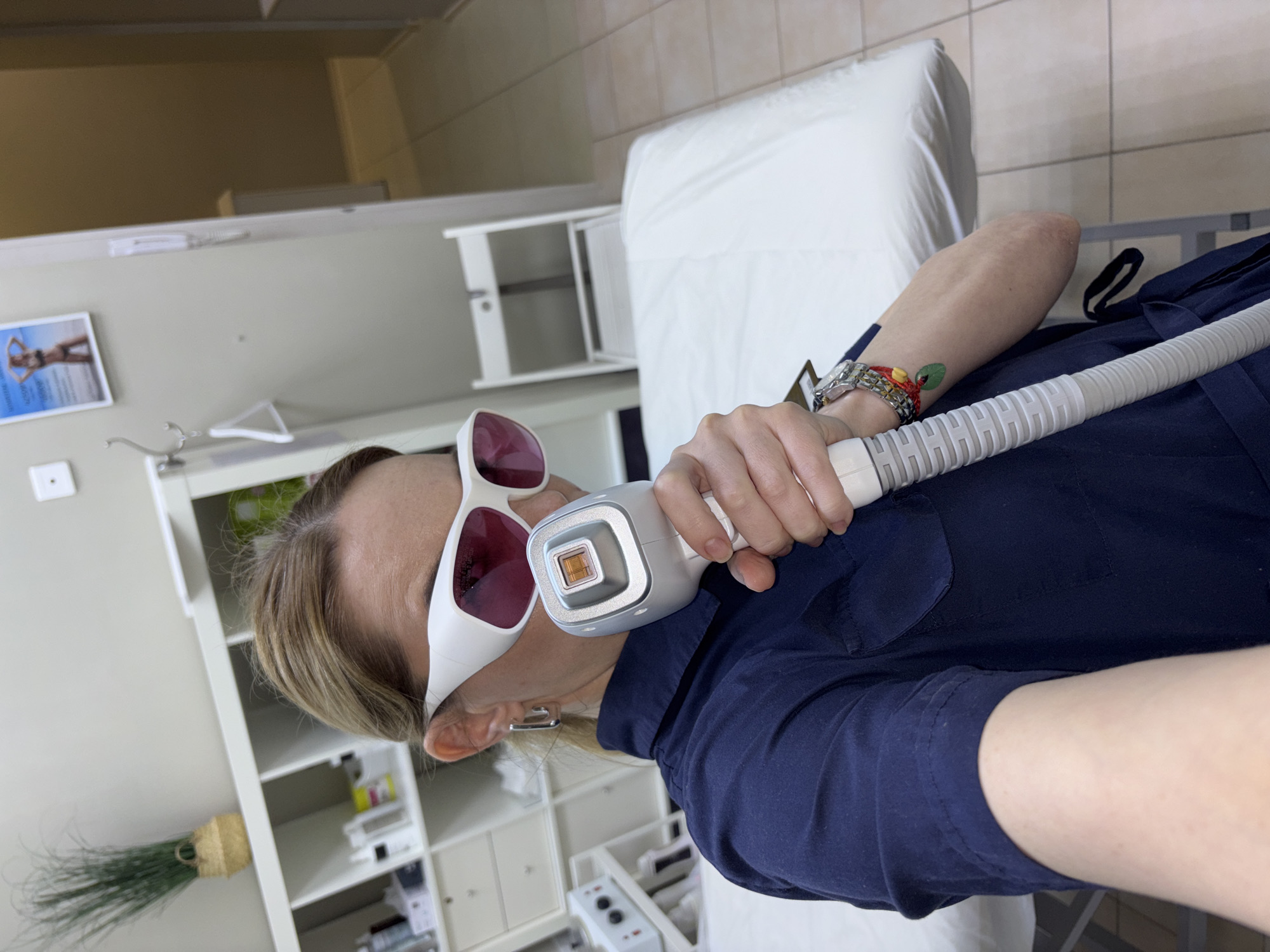 The image shows a woman in what appears to be a medical or cosmetic clinic. She is wearing white protective eyewear with dark pink lenses and holding a handheld laser device with a ribbed hose. She is wearing a blue top.
The background includes a white medical examination bed, a white shelving unit with various items, and tiled walls. A framed image of a woman in a bikini hangs on the wall.
Based on the instructions, the location is Vsevolozhsk, Russia.