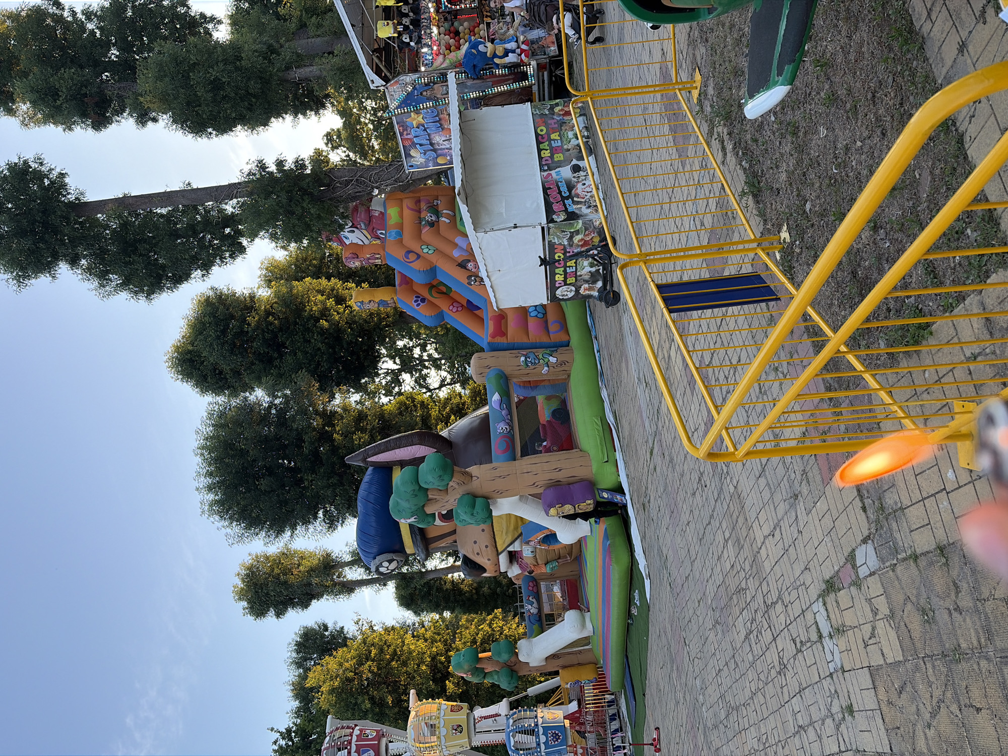 The image depicts an outdoor amusement area with various attractions under a clear sky with scattered clouds. In the foreground, a yellow metal fence lines a paved area with interlocking brick patterns. To the left, part of a red scooter is visible. Dominating the center of the image is a large inflatable obstacle course featuring slides, climbing areas, and characters. Adjacent to this is a Ferris wheel with colorful carriages. Further to the right, there is a game booth with prizes, including stuffed animals, and a sign that reads "STAR WARS". The area is surrounded by tall trees. A white tent with colorful graphics advertising "DRAGON BREATH ICE CREAM" is situated in front of the game booth. A few people are present in the scene, including a person operating the inflatable structure, a woman with a stroller, and a man standing near the game booth. The lighting suggests it is late afternoon or early evening.