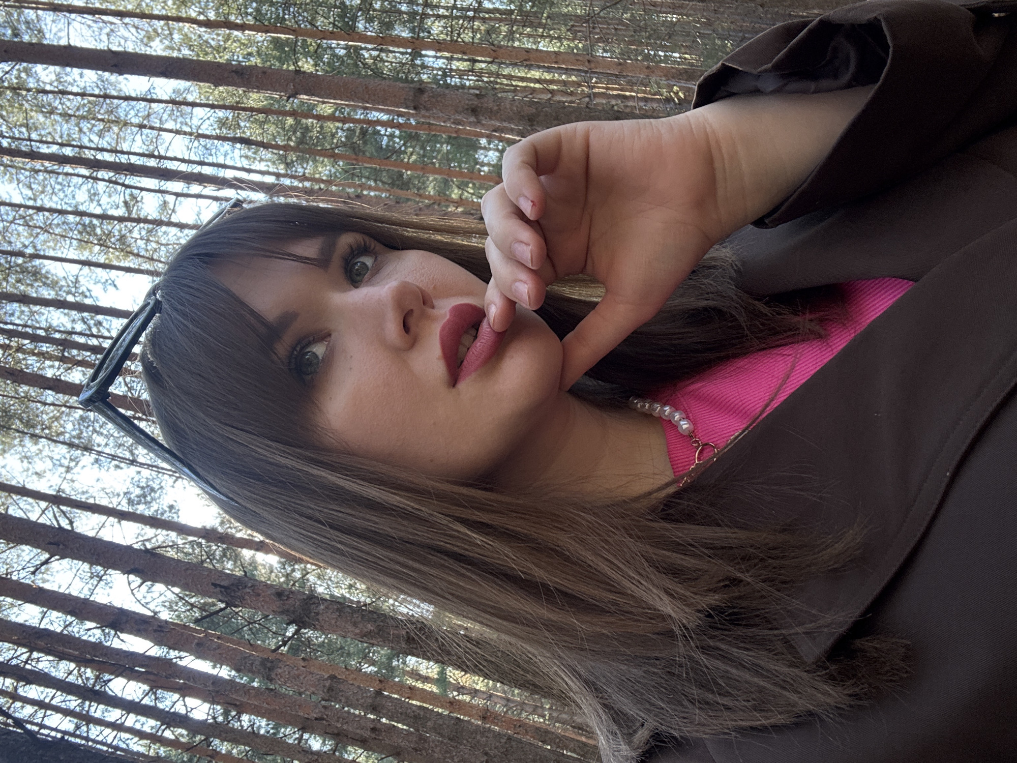 A portrait-style image captures a young woman from an upward, tilted perspective, set against a forest background. She has long, straight brown hair with bangs, and striking green eyes. Her lips are painted with bright pink lipstick. Sunglasses are perched on top of her head. She wears a bright pink top underneath a dark brown or black coat, and a pearl necklace is visible around her neck. Her left hand is raised, with fingers gently touching her chin and lower lip in a posed or thoughtful gesture. The background consists of numerous tall, slender trees, likely pines, with their trunks forming a dense, repetitive pattern against a bright, clear sky, indicating sunny daytime conditions. The setting is a forest in Chaikovsky, Russia. There is no visible text in the image.