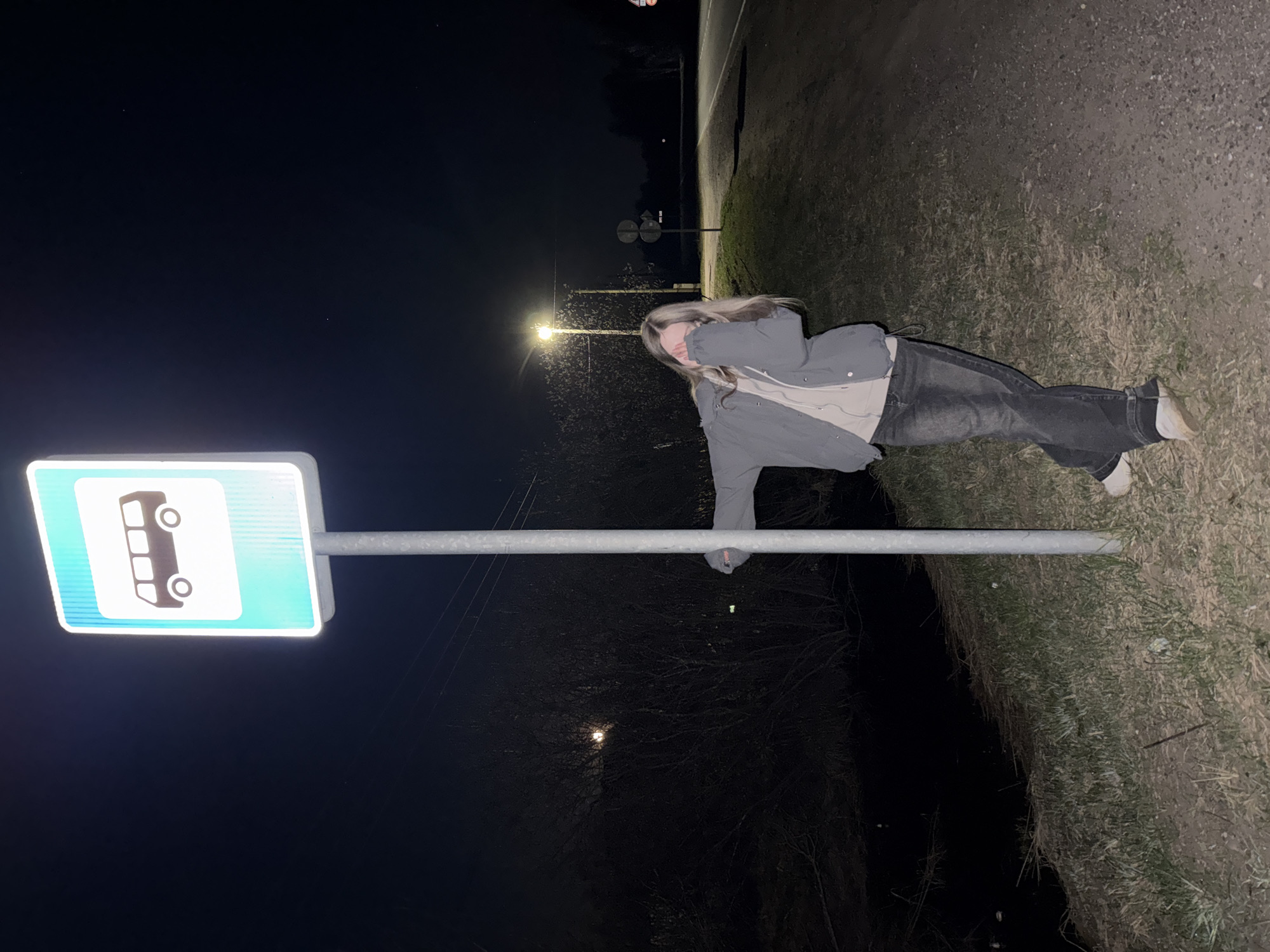 At night in an unidentified location in Russia, a woman poses in front of a bus stop sign. The woman has blonde hair and wears a gray jacket over a tan undershirt, along with dark jeans and white shoes. The bus stop sign is illuminated, showing a white bus symbol on a blue background. The surrounding environment includes dark foliage, a road, and distant streetlights.