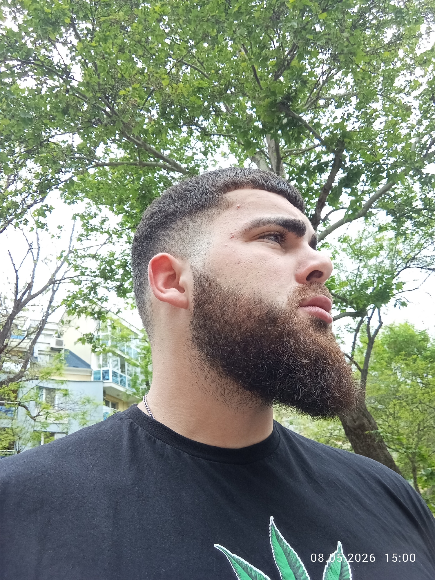 A medium shot captures a man with a thick, well-groomed brown beard and a fade haircut, looking upwards to the right. He is wearing a black t-shirt with a graphic design of green leaves and a silver necklace. The image also contains a date stamp with the date 08.05.2026 and the time 15:00.
The background consists of green trees and vegetation. In the distance, a building with blue and white accents is visible. This building's architectural style is consistent with buildings found in Plovdiv, Bulgaria. The surrounding environment suggests a park or garden setting, which is commonly found in urban areas of Plovdiv.