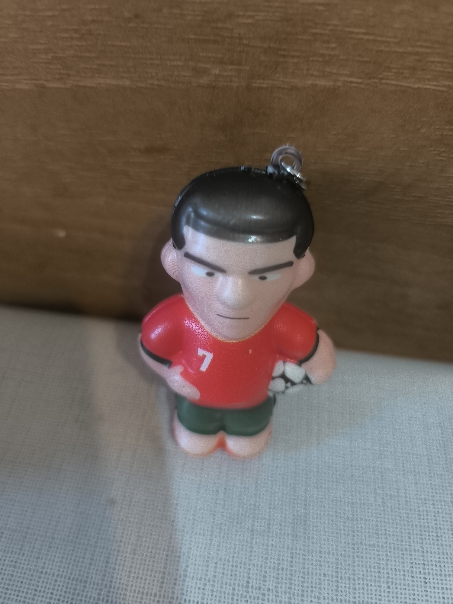 The image displays a small, stylized figurine of a male soccer player, likely serving as a keychain due to the metallic loop attached to the top of its head. The figure is cartoonish in design, featuring black hair, prominent black eyebrows, and a simple line for a mouth, conveying a neutral or slightly stern expression. It wears a bright red short-sleeved shirt with black cuffs, bearing the visible white number "7" on its chest. Dark green shorts complete the uniform. A black and white soccer ball is held securely under the figure's left arm. The figurine stands on a light-colored, subtly textured surface, possibly a fabric with a grid-like weave. Behind the figure, a blurred, warm brown wooden surface is visible, suggesting an indoor setting. The lighting is even and bright, but the specific time of day is not indicated. No particular activity is depicted beyond the static pose of the figurine. This object is located in Yerevan, Armenia.