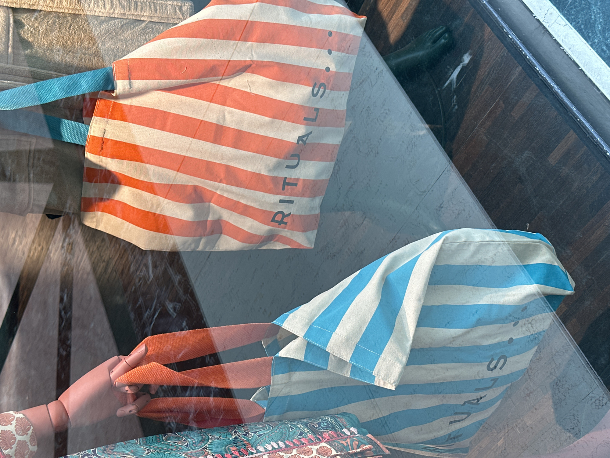 The image shows two striped tote bags in a window display. One bag has orange and white stripes with blue handles, and the other has blue and white stripes with orange handles. Both bags have the word "RITUALS" printed vertically on them. A mannequin hand and a green statue are visible through the window. The background includes a wooden floor and a textured surface. The city and country are not provided in the image.