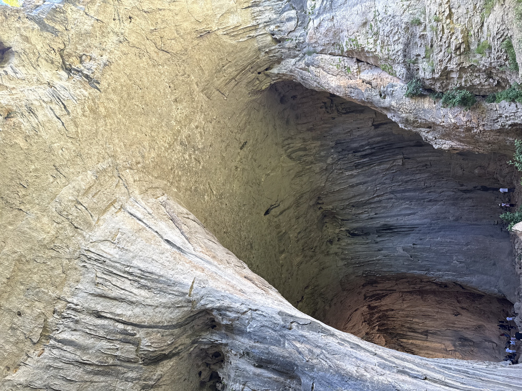 This image showcases the Devetashka Cave, located near the city of Karlukovo in Bulgaria. The cave is a large natural cavern with a wide entrance, featuring layers of stratified rock. A group of people is visible near the cave entrance. Green vegetation grows on the rock surfaces. The cave has a varied texture, marked by cracks and layered sedimentary rocks. Sunlight penetrates the cave, illuminating its interior.