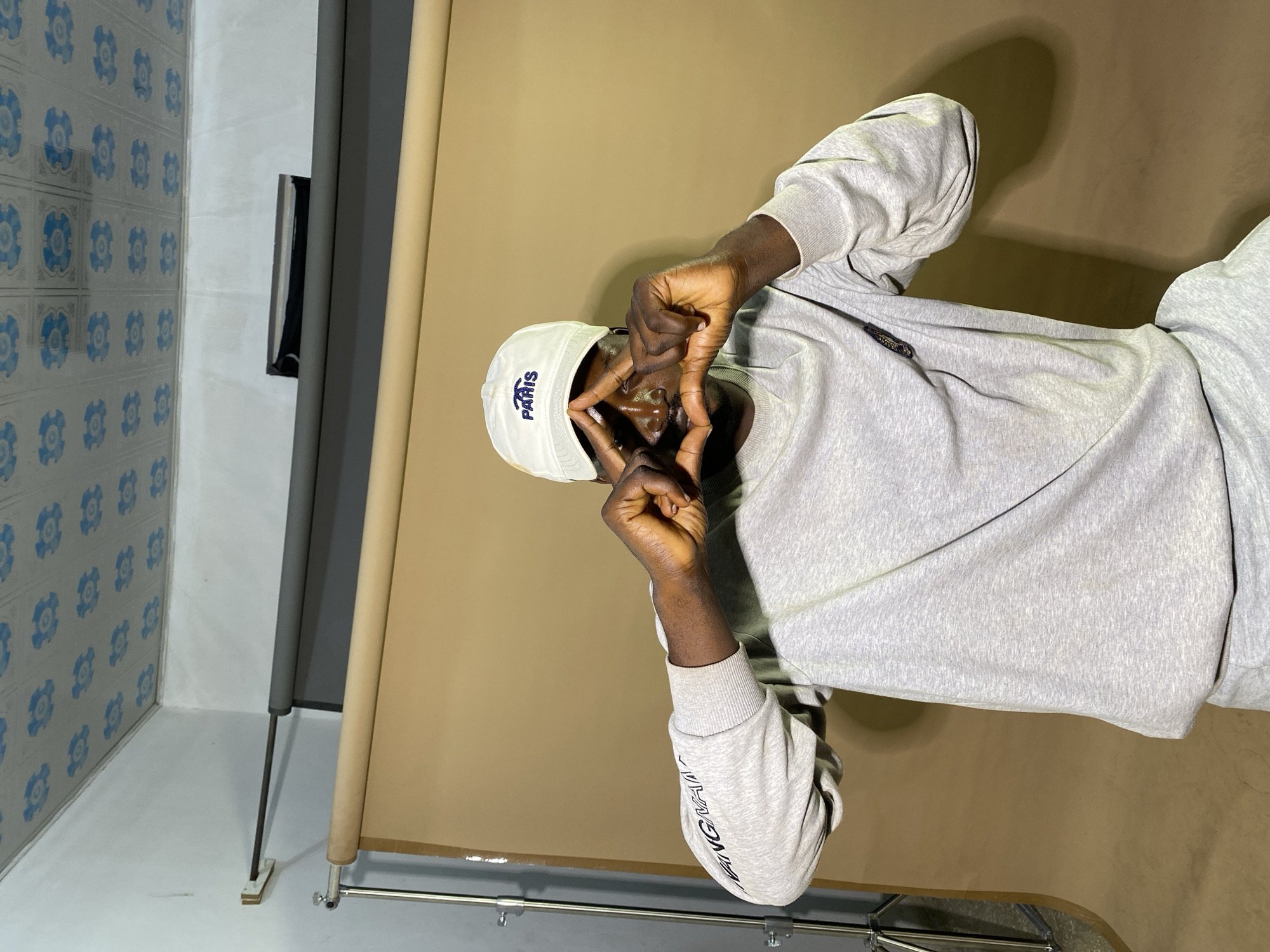 The image depicts a young Black man wearing a white baseball cap with "PARIS" written on it, and a light grey sweatshirt. He is seated and holding his hands up to his face, forming a triangle shape with his fingers in front of his eyes. He is positioned in what appears to be a makeshift studio setting, with a large, tan backdrop behind him. The ceiling of the room has a patterned finish with blue floral designs. A lighting stand is visible to the left of the frame. The man's posture and the studio setup suggest a portrait or photoshoot. The specific location within the city of Gura Top, Nigeria is not determinable from the visual information provided.