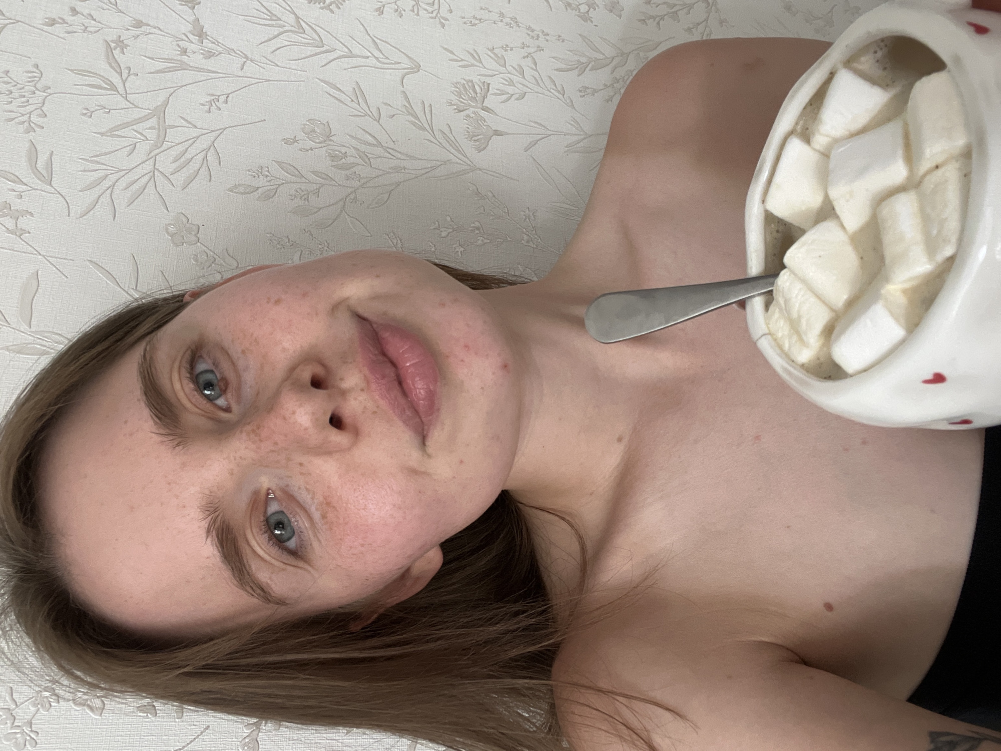 A young Caucasian woman with light brown hair and blue eyes is looking into the camera, holding a mug of hot chocolate with marshmallows. She is bare-shouldered and smiling slightly. The background is a white wallpaper with a floral pattern. The image appears to be taken indoors. No specific location in Chaikovsky, Russia is identifiable from the image.