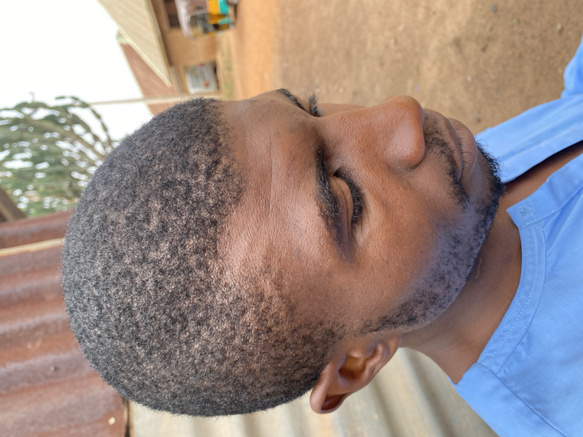 A close-up headshot depicts a Black male with short, dark hair and a trimmed beard. His eyes are closed, and his skin tone is medium brown. He is wearing a light blue collared shirt. The background is blurred, but includes corrugated metal and foliage. The image was likely taken outdoors. Based on the instructions, the location is Kuru Karama, Nigeria.