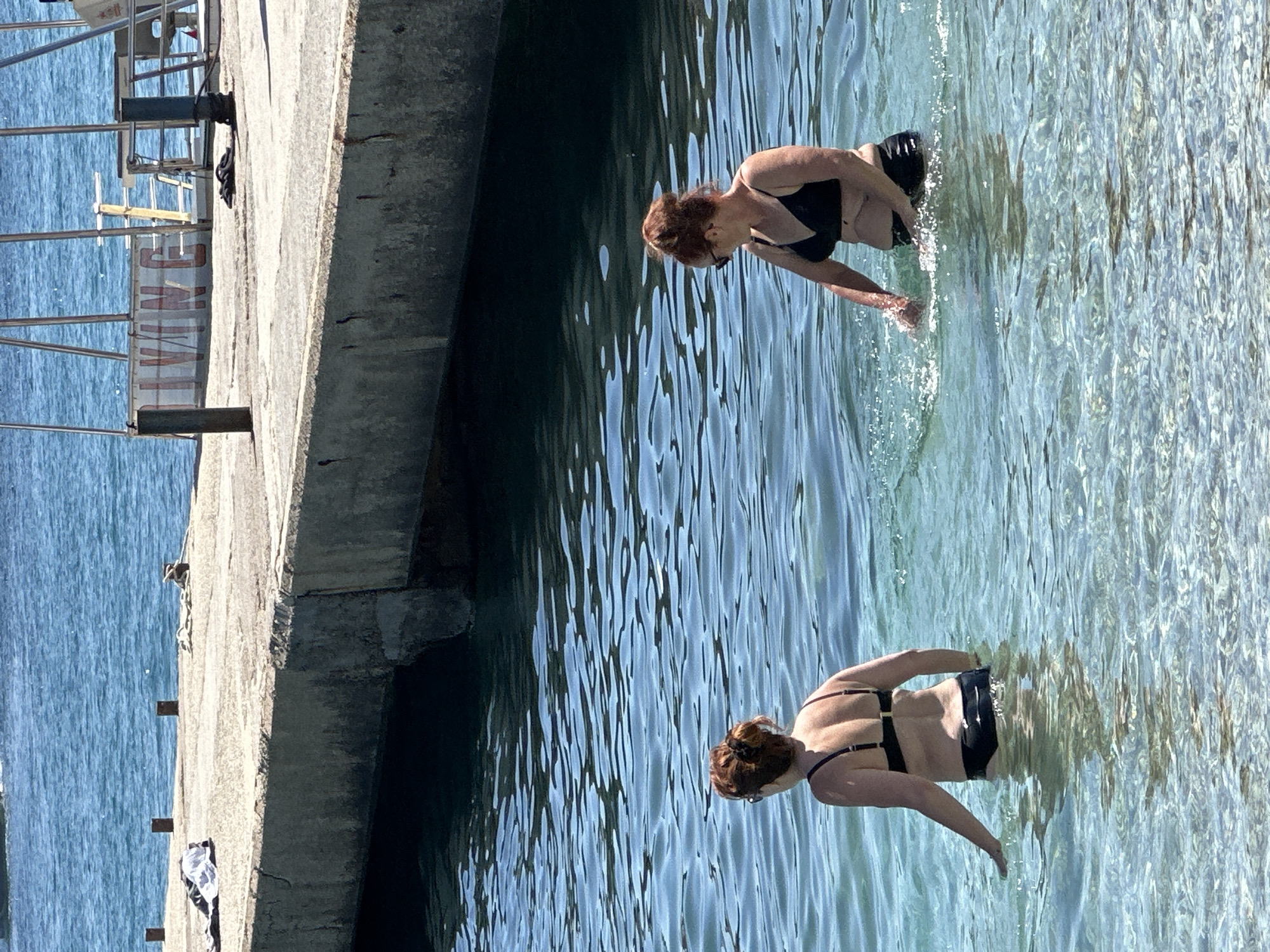 Two women are standing in clear, blue water near a concrete pier. The woman on the left has her back to the camera, wearing a black bikini. The woman on the right is facing partially to the left, also in a black bikini, and is splashing water with her hands. The pier appears to be part of a diving platform, with metal structures and a sign reading "DIVING" visible in the background. The water is shallow enough that the women's legs are visible. The scene is set in a sunny, outdoor environment, likely a coastal area.