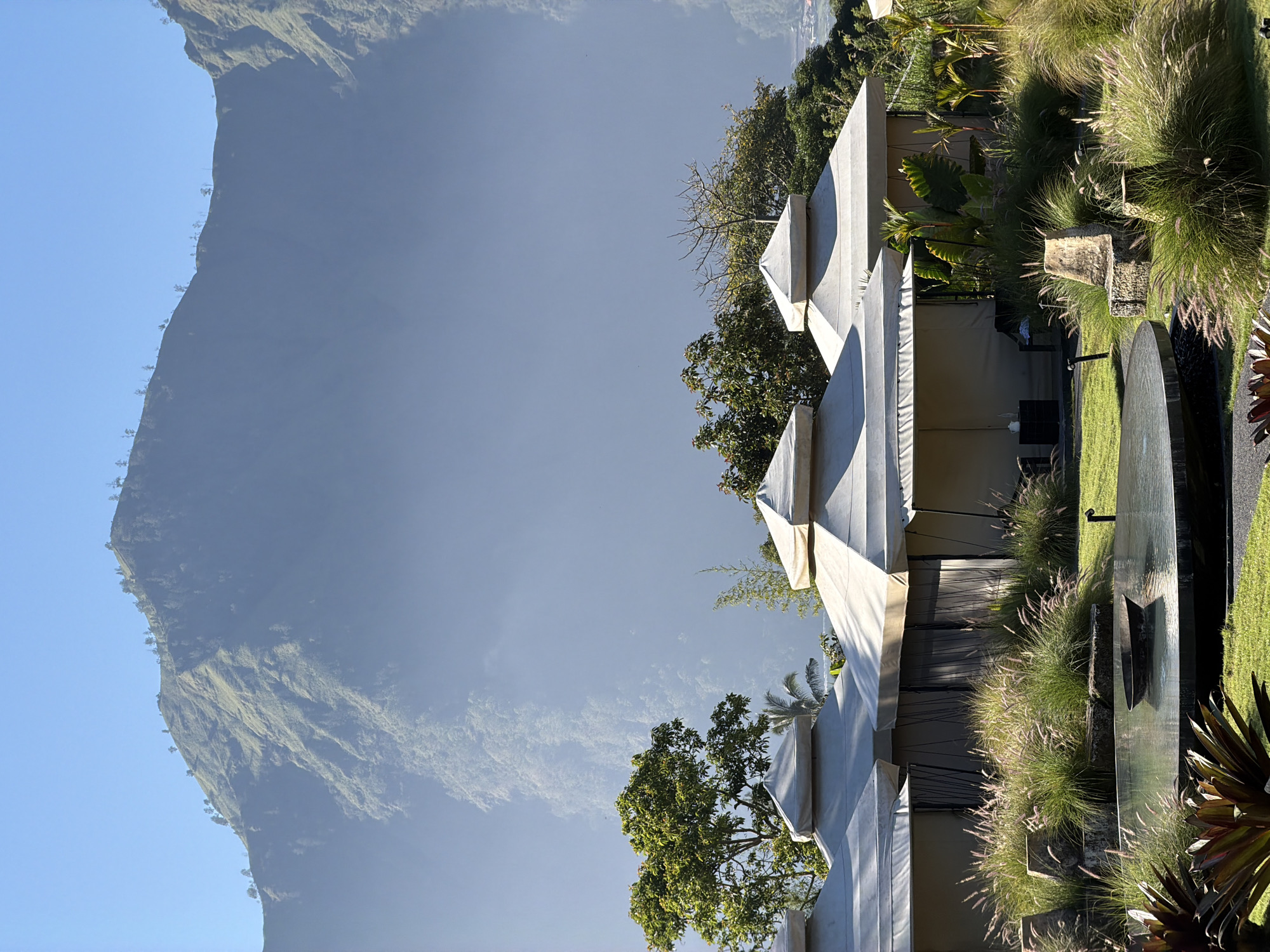 The image depicts a serene outdoor scene at a resort in Kintamani, Indonesia. In the foreground, a circular water feature with a dark metal basin is surrounded by lush green grass and ornamental plants. Several canvas tents with peaked roofs are visible in the middle ground, set amidst a landscape of tall grasses and tropical foliage. Behind the tents, a dense forest covers the slopes of a large, misty mountain that dominates the background under a clear blue sky. The scene suggests a tranquil and picturesque environment, typical of a high-altitude retreat.