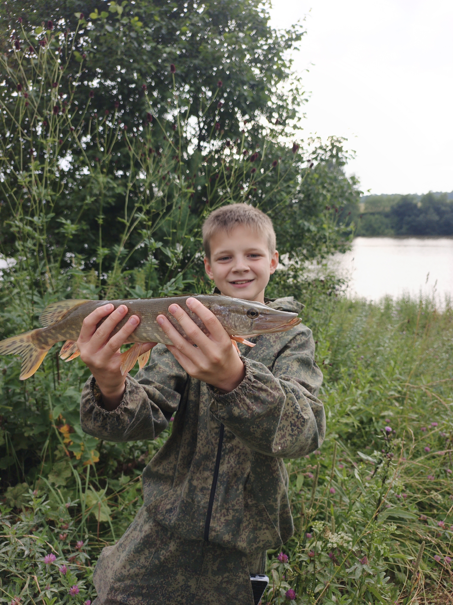 A young boy, appearing to be around 10 years old, stands in a natural setting near a body of water. He is wearing a camouflage jacket and proudly holds a freshly caught fish horizontally in front of him with both hands. The fish is long and slender with scales and fins visible. The boy has a smile on his face, suggesting he is pleased with his catch.
The background consists of lush green foliage, including trees and tall grasses, with small purple flowers scattered among the greenery. In the distance, a calm body of water, likely a river or lake, is visible, bordered by more trees. The sky is overcast. The overall scene depicts a moment of successful fishing in a rural or semi-rural environment, possibly on the outskirts of Urtiushka, Russia, given the visual cues of the landscape.