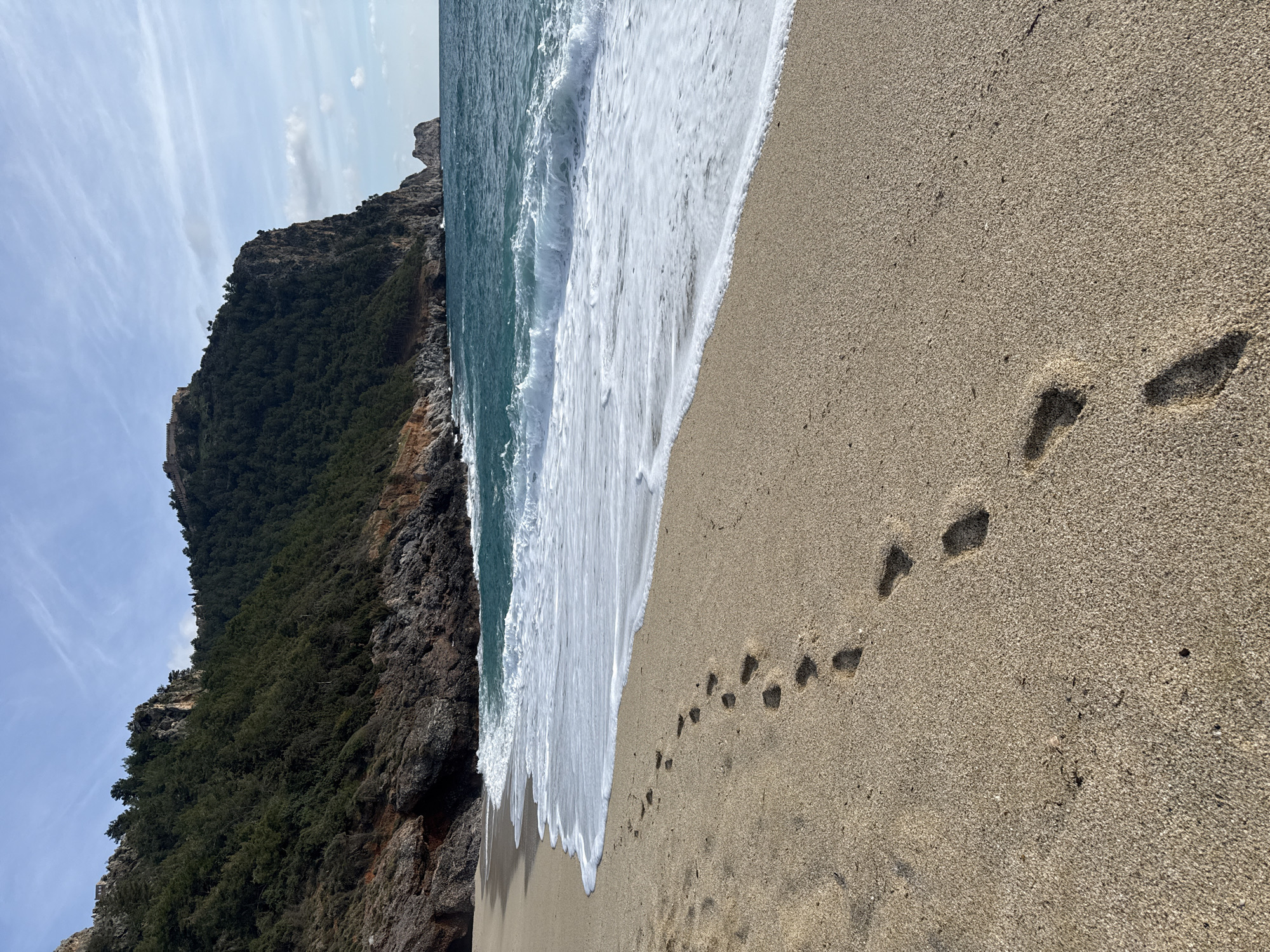 The image displays a coastal scene in Alanya, Turkey, specifically a beach segment at the base of the Alanya Peninsula. The foreground features light beige sand, where a series of approximately ten human footprints extends diagonally from the lower left towards the center right, parallel to the water's edge. The sand appears damp where it meets the water. The midground consists of the turquoise-blue Mediterranean Sea, characterized by multiple white-crested waves actively breaking and receding, forming a continuous line of white foam along the shoreline.
The background is dominated by a large, steep promontory, densely covered in dark green coniferous trees. Sections of exposed reddish-brown and dark grey rock face are visible through the vegetation. At the apex of this promontory, historical fortifications, identifiable as parts of Alanya Castle, are discernible. In the far right distance, a solitary rocky islet is present off the coast. The sky above is partially cloudy, displaying light blue hues with scattered wispy white cloud formations. No individuals are visible within the frame; the footprints are the sole evidence of human presence or activity.