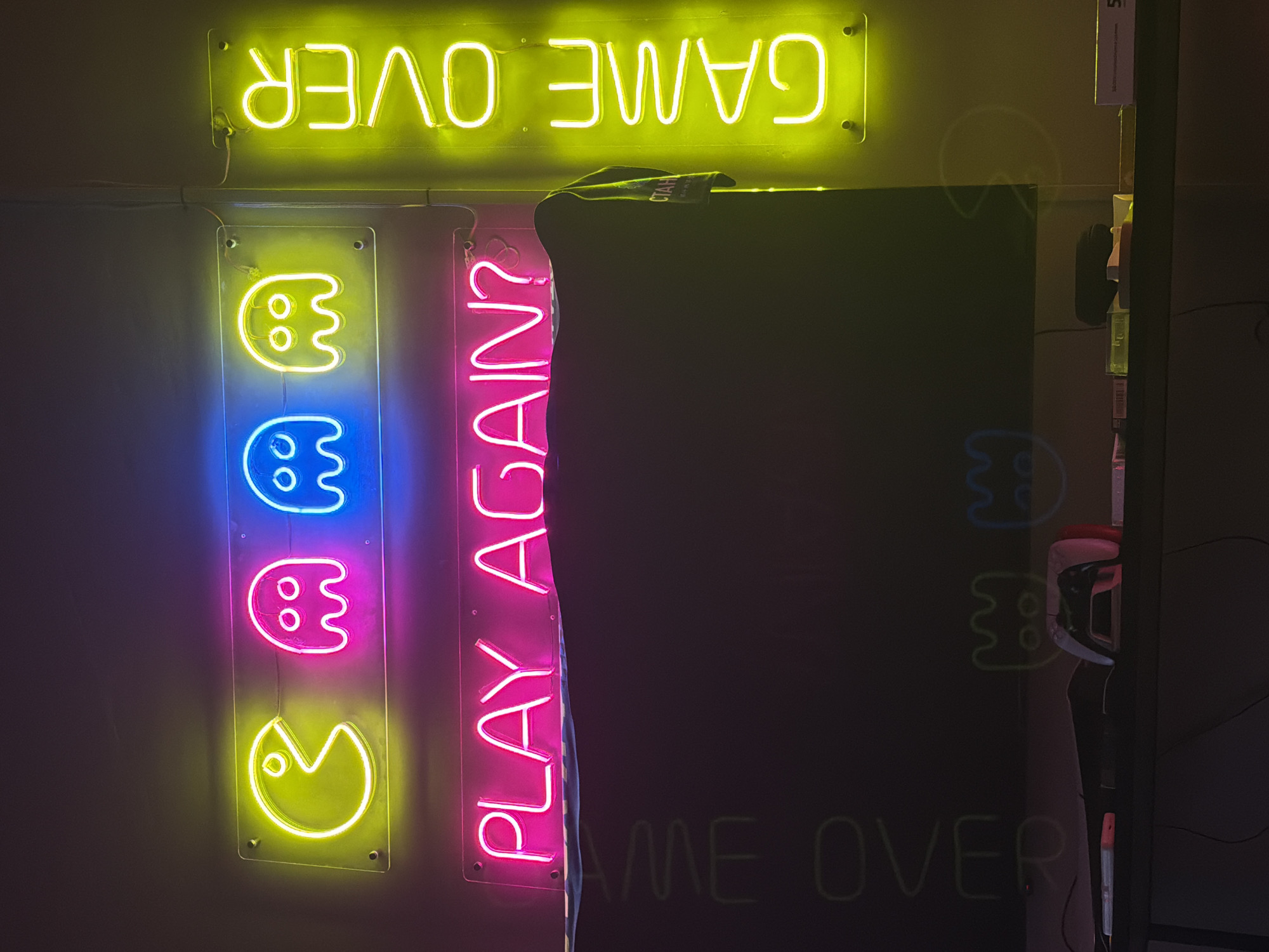 The image shows a section of wall art consisting of neon signs. The top sign reads "GAME OVER" in bright yellow, mounted horizontally. Below, a vertical sign displays a Pac-Man and three ghosts, each in a different color, going from yellow at the bottom to pink, then blue, and then yellow at the top. Next to it is another vertical sign that reads "PLAY AGAIN?" in bright pink neon. To the right, a black screen with faint neon outlines of ghosts and "GAME OVER" beneath. In the background, part of an arcade machine and other assorted objects are visible. There is no specific evidence to indicate that the image was taken in Izhevsk, Russia.